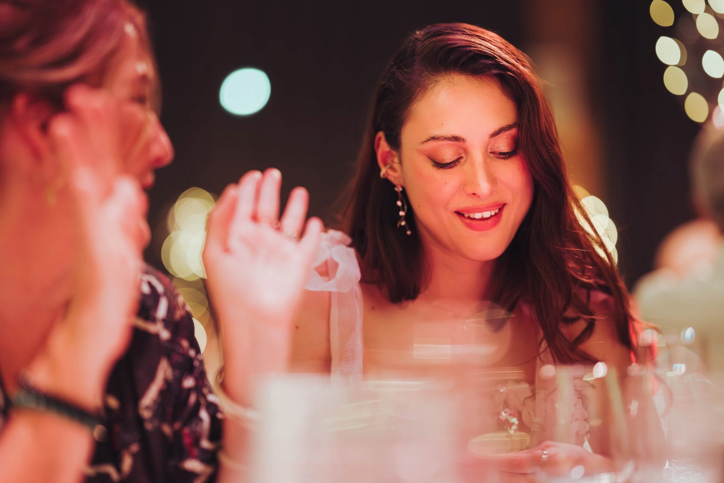 Deux femmes souriantes assises à une table au Mas Tolosa, proche de Toulouse, en Occitanie, lors d'une soirée festive de mariage avec des lumières floues en arrière-plan