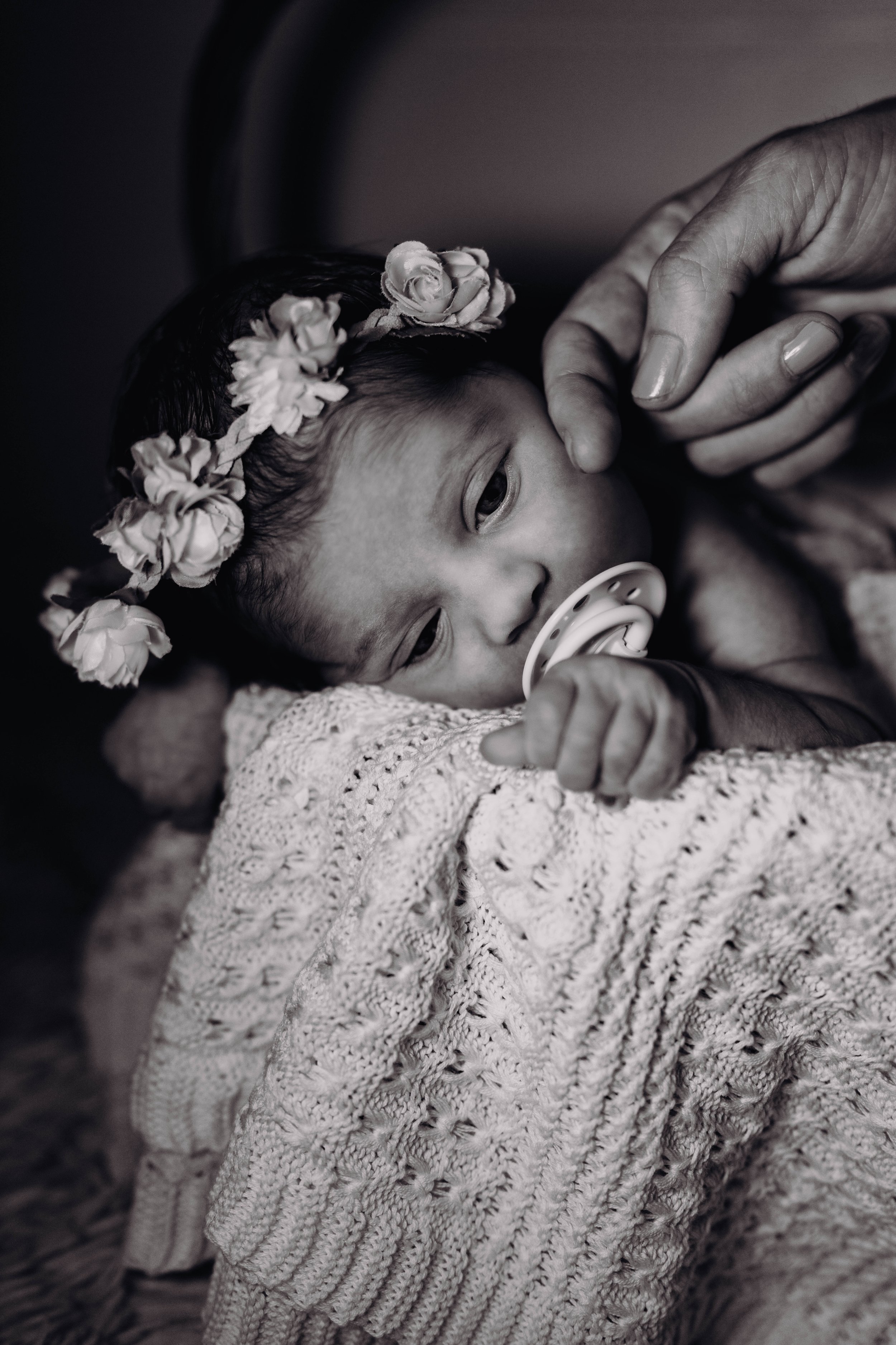 Une jeune enfant avec un headband de fleurs, allongée sur une couverture, suçant un tétine, tenue doucement par une main.
