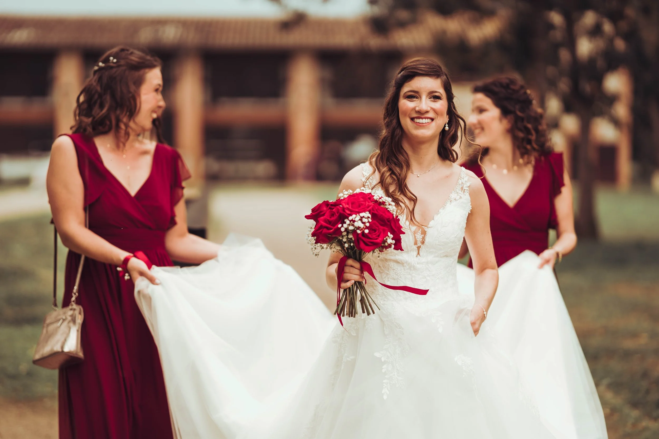 Une mariée souriante accompagnée de deux femmes en robes rouges lors d'un mariage à l'extérieur.
