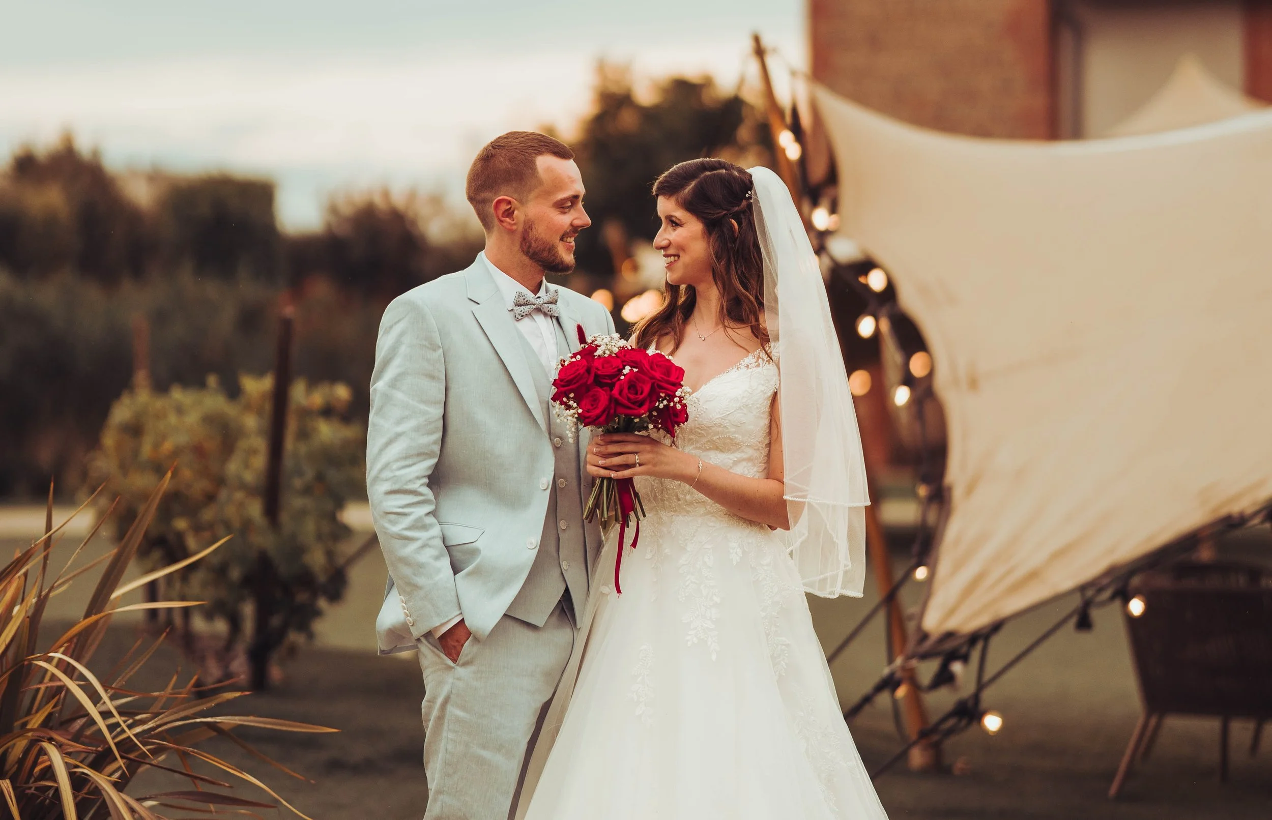 Un couple de mariés souriant lors de leur mariage en extérieur au Mas Tolosa proche de Toulouse, la mariée tenant un bouquet de roses rouges et portant une robe blanche, l'homme en costume clair avec une cravate en nœud papillon.