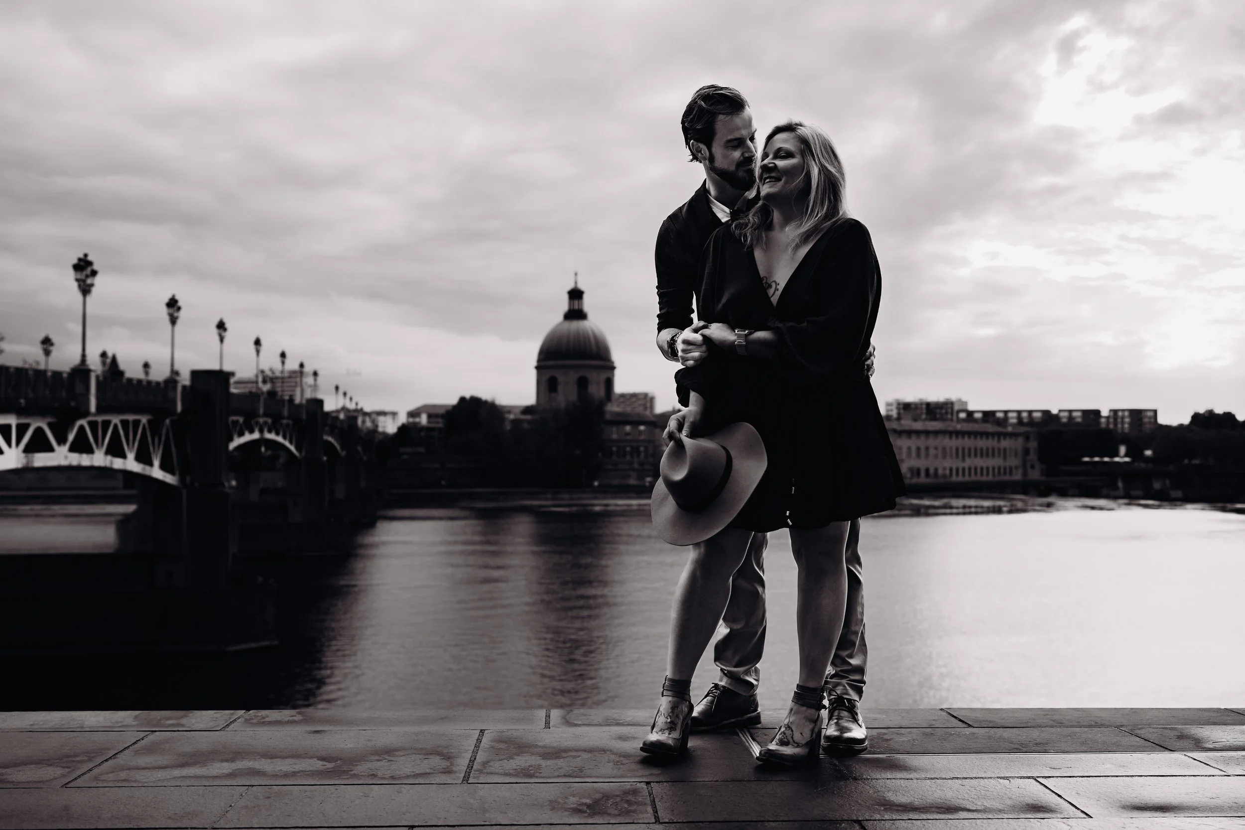 Un couple en noir et blanc se tient près d'une rivière, avec un pont et un bâtiment avec un dôme en arrière-plan, un jour nuageux.