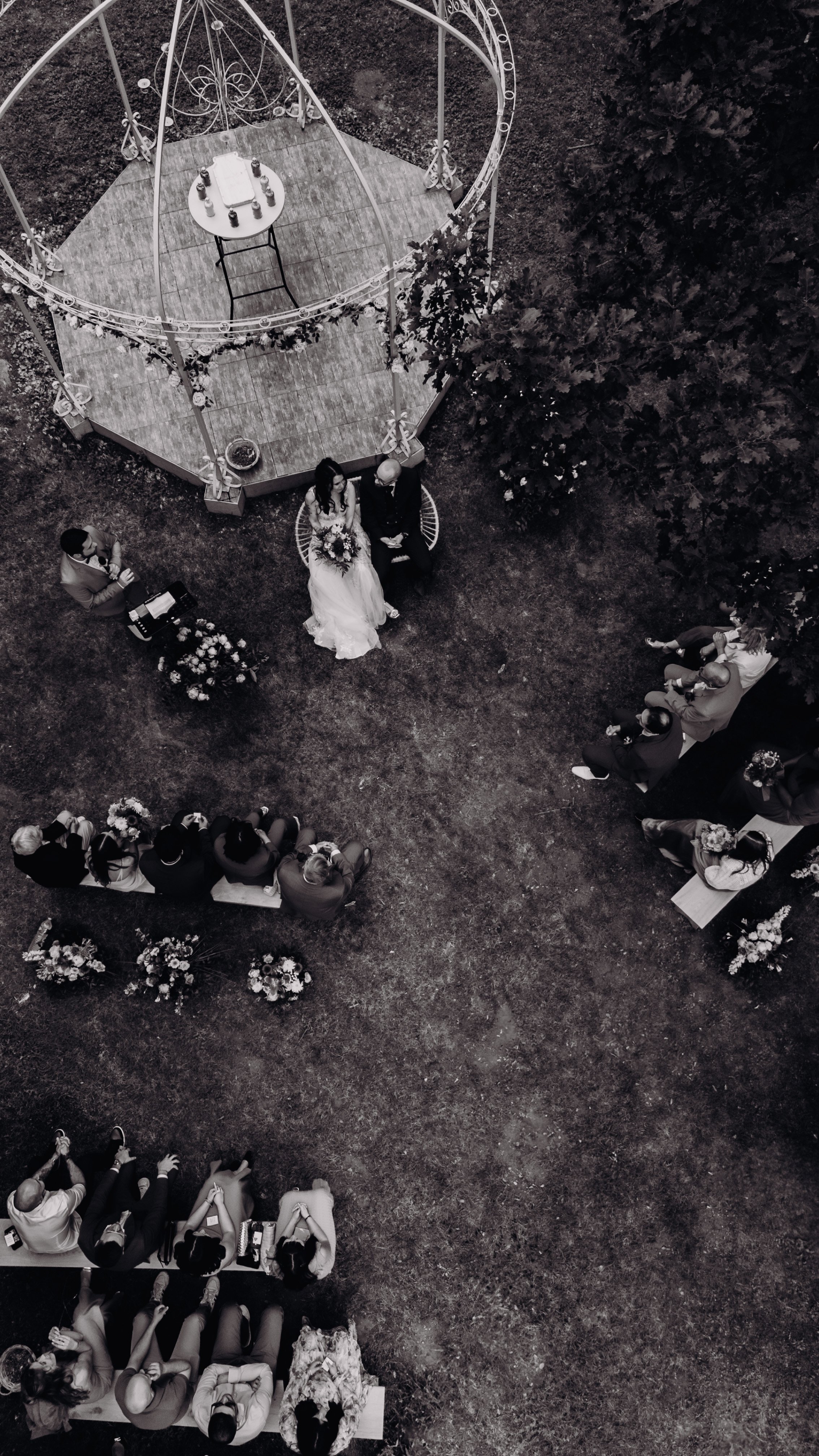 Cérémonie de mariage en plein air au domaine de Lalanne, vue de drone, avec les mariés assis sous une arche, entourés par des invités assis sur des bancs, dans un cadre naturel