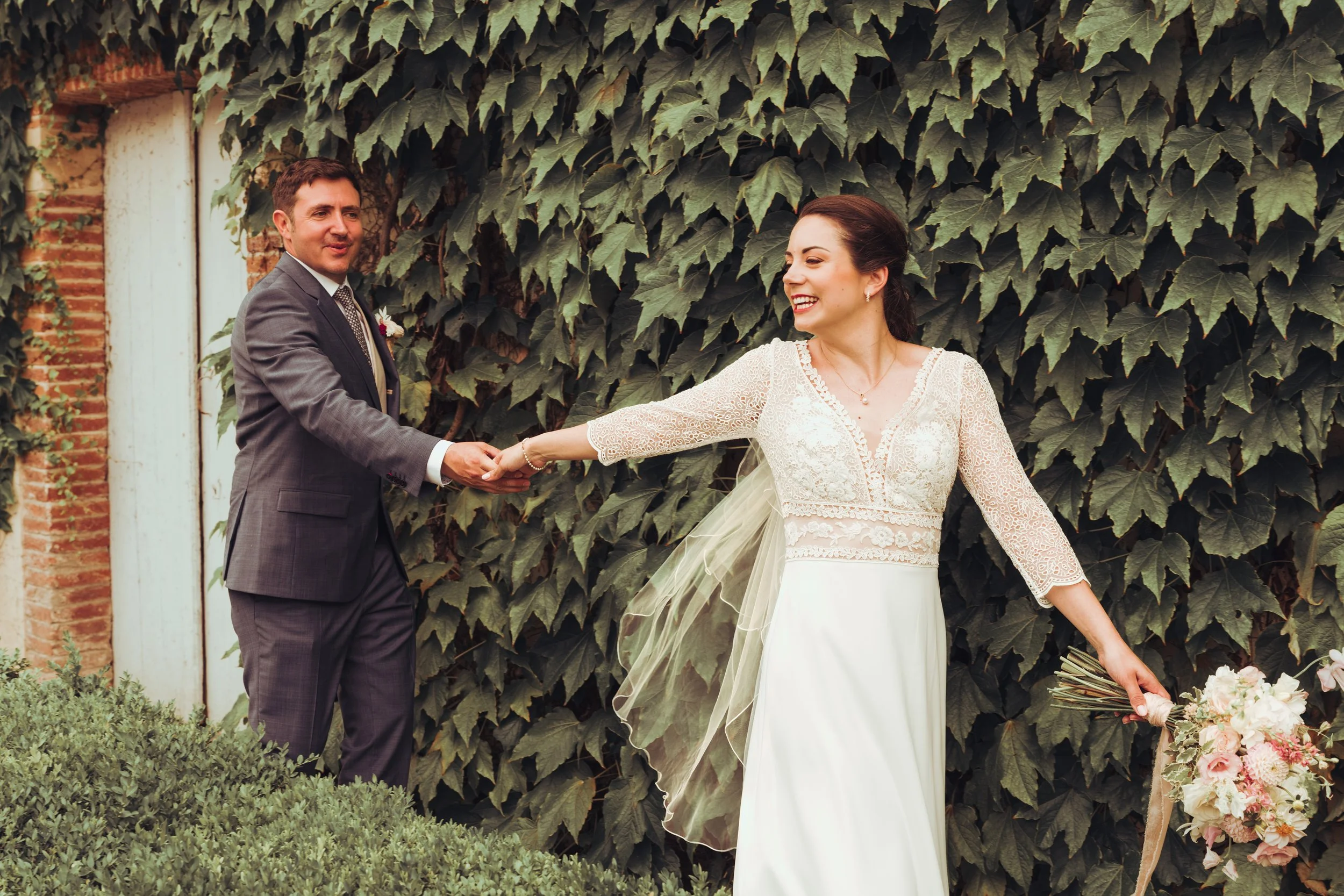Un couple de mariés se tenant la main au domaine de Gailhaguet proche de Toulouse, en Occitanie, la femme souriante en robe de mariage blanche, la scène en plein air devant une haie verte dense.