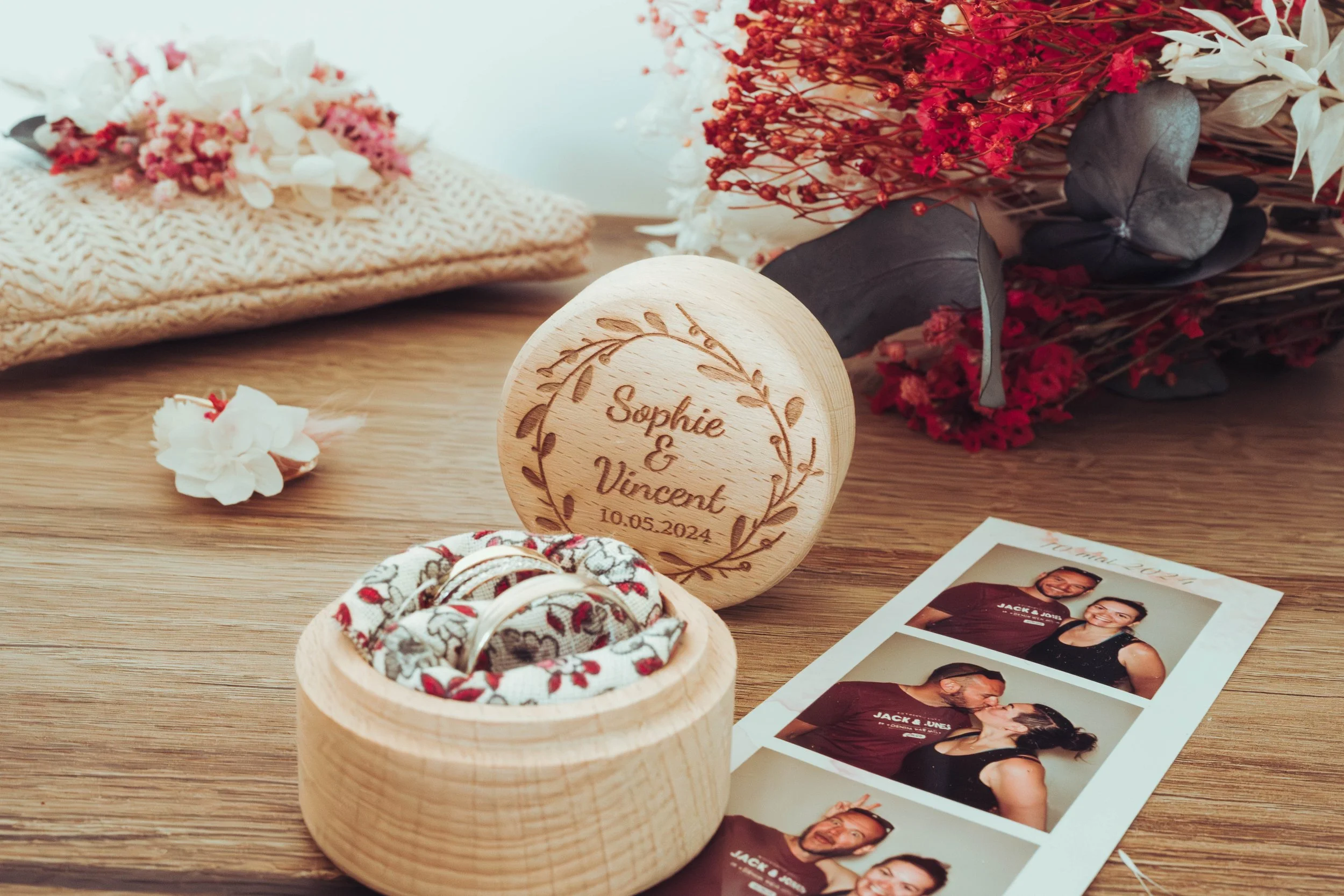 Flatlay réalisé dans un mariage à Grenade proche de Toulouse. Une boîte à souvenirs en bois gravée avec les noms Sophie et Vincent entourée de fleurs, d'une photo de couple, et de plusieurs alliances, sur une table en bois.