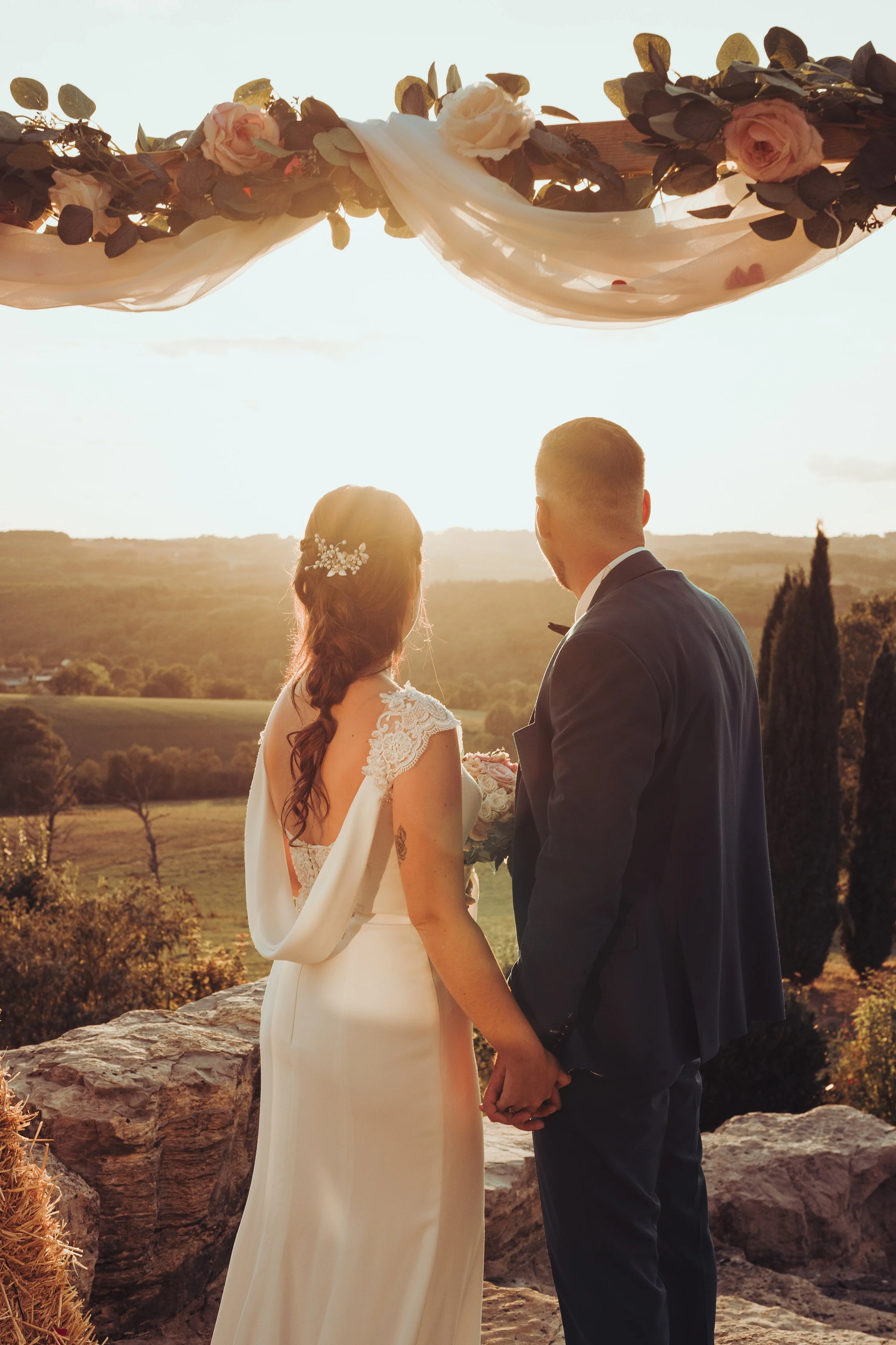 Un couple de mariés se tenant la main, regardant un coucher de soleil sous leur arche de mariage fleurie, devant les vallons de région Toulousaine. Vue rurale avec des collines et des arbres faisant penser à la Toscane. 