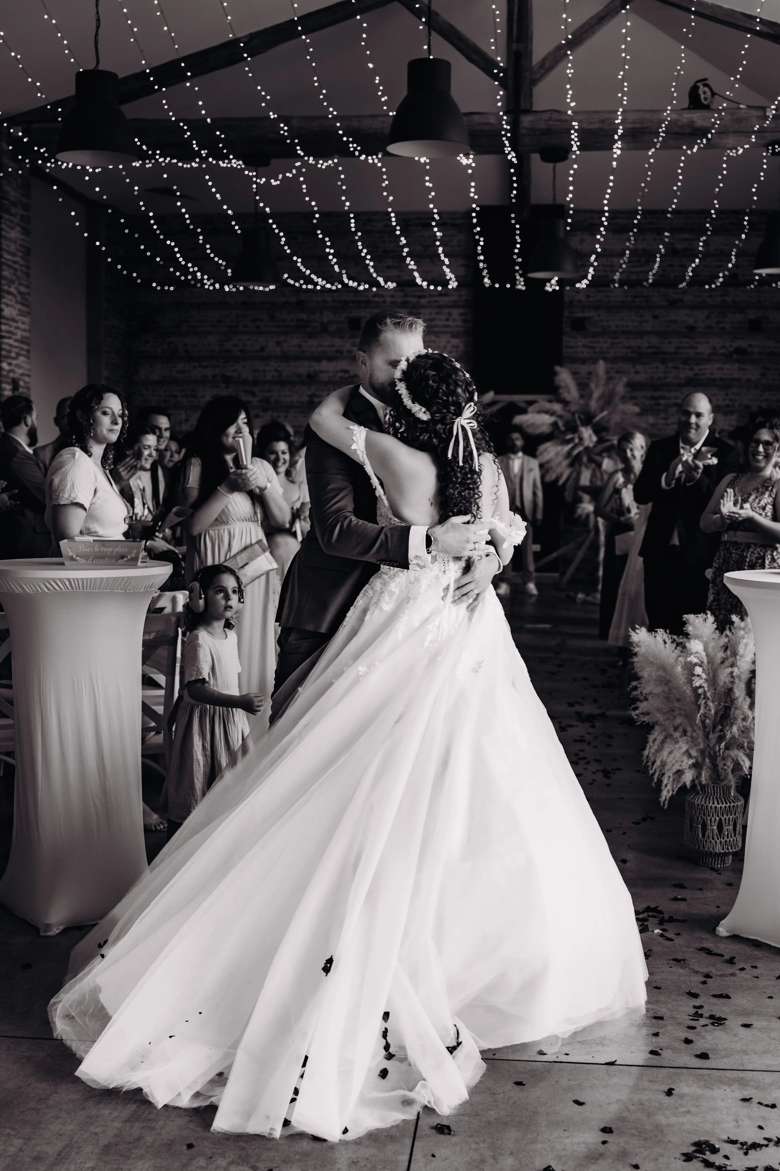 Un couple de mariés en train de danser lors de leur mariage à la métairie du Fusté proche de Toulouse, en Occitanie, entouré d'amis et de famille dans une salle décorée avec des guirlandes lumineuses.