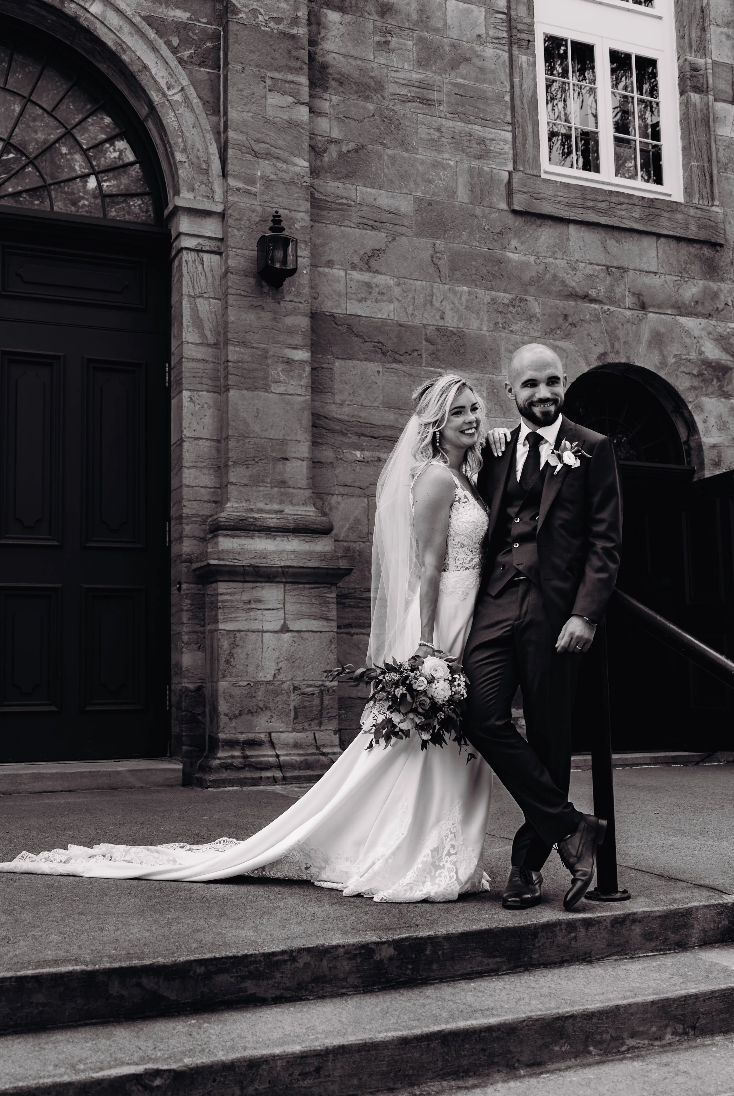 Un couple marié, un homme et une femme, posant devant un bâtiment en pierre, en tenue de mariage. La femme tient un bouquet de fleurs et porte une robe de mariage avec voiles, l'homme porte un costume noir avec une boutonnière.