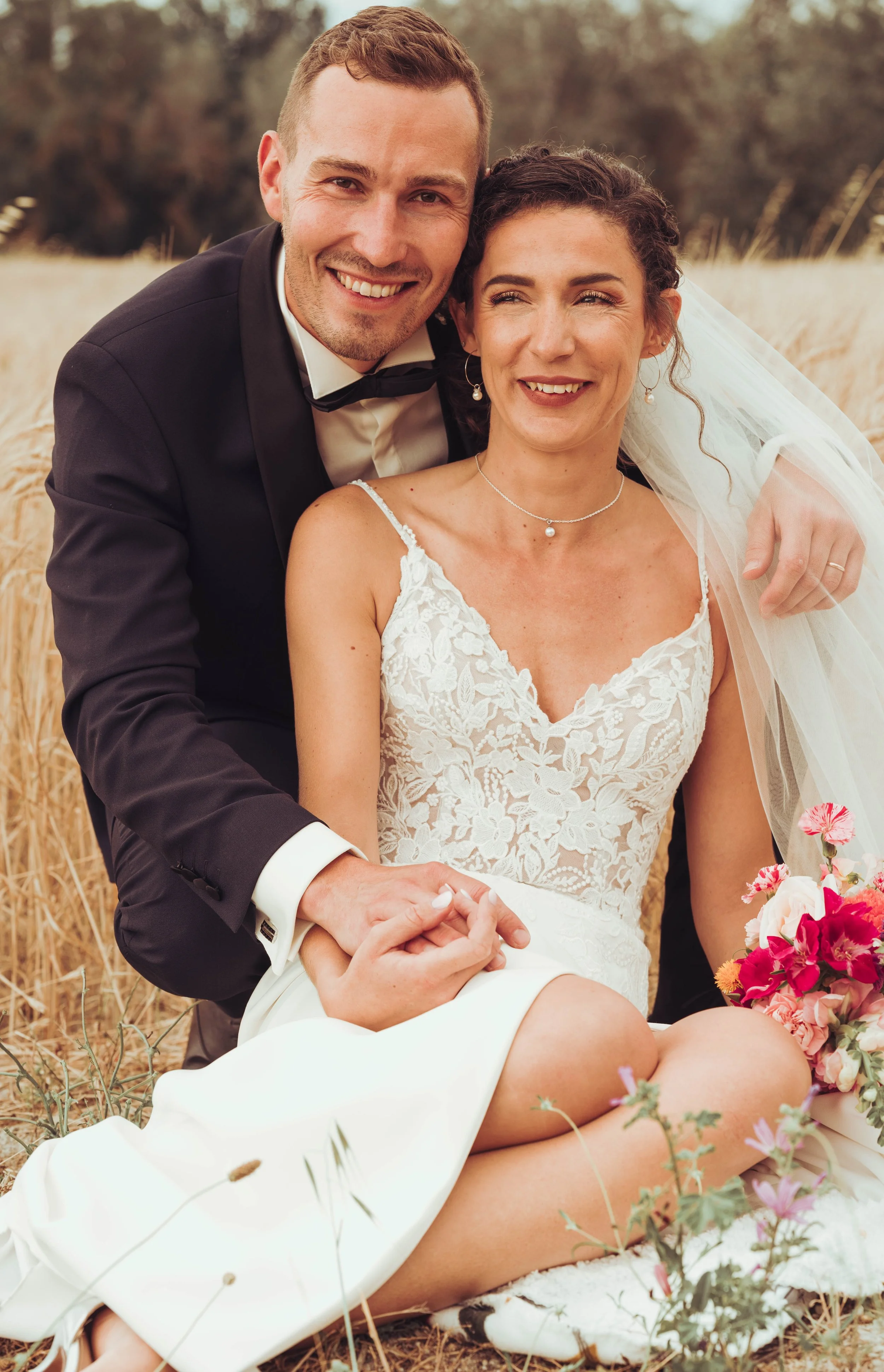Un couple en mariage assis dans un champ de blé proche de Béziers, en Occitanie, la femme en robe de mariée en dentelle et le homme en costume noir, tenant la main de la mariée, tous deux souriants.