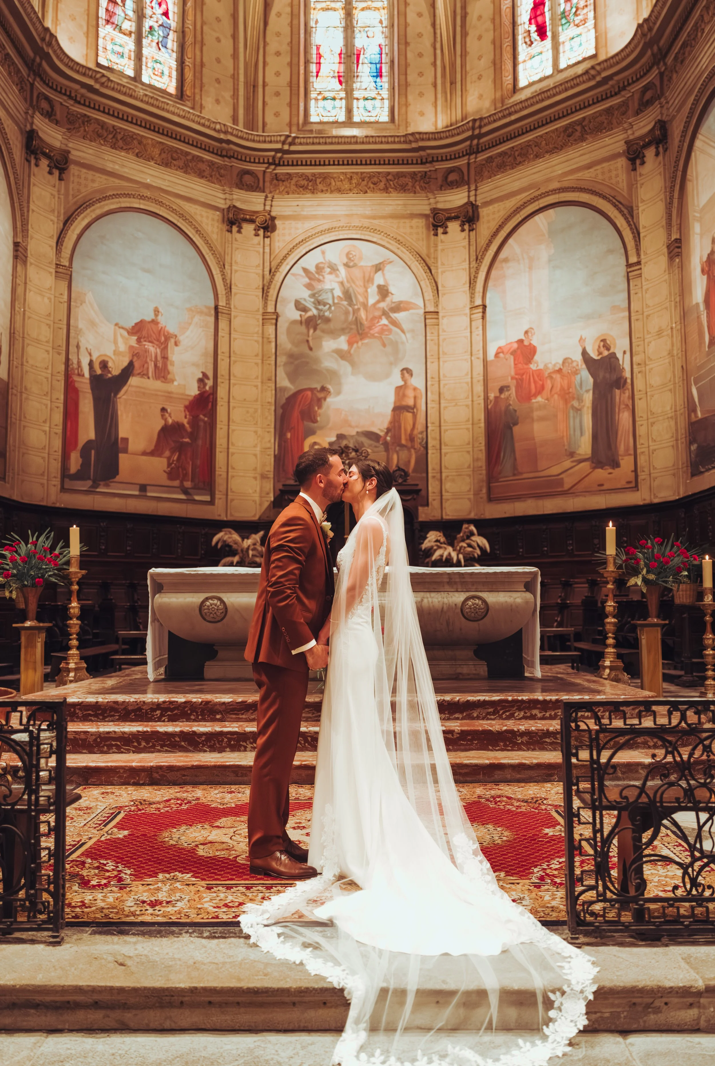 Un couple de mariés s'embrassant dans une cathédrale proche de Toulouse, avec un décor somptueux, des peintures religieuses sur les murs et des vitraux colorés au plafond.