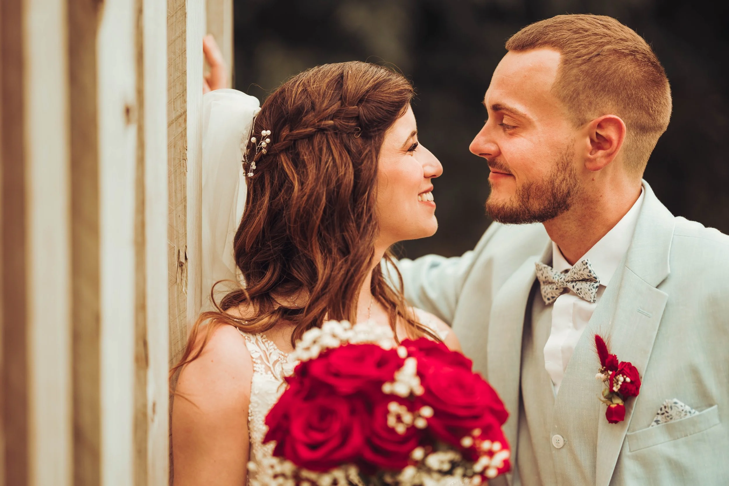 Un couple de mariés au Mas Tolosa proche de Toulouse, en Occitanie, souriant, se tenant face à face, devant un mur en bois, la femme tient un bouquet de roses rouges et la mariée porte une robe blanche avec des détails en dentelle.