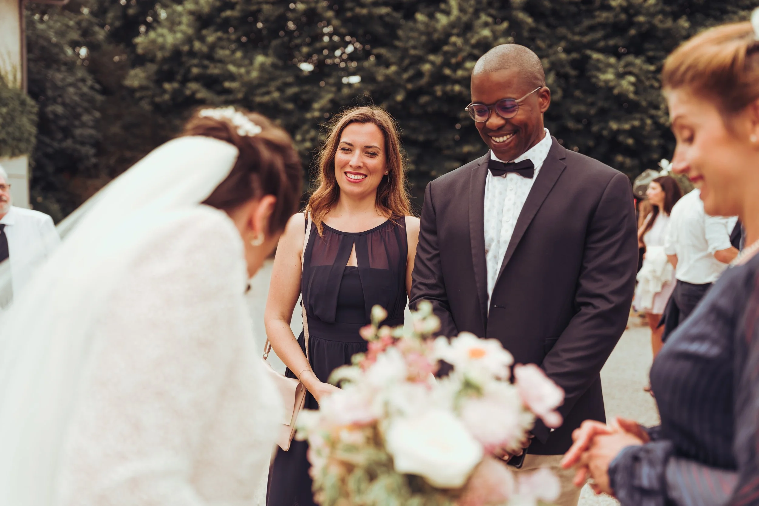 Un couple de mariés au mariage, entouré d'amis qui sourient, lors d'une cérémonie en plein air avec des arbres en arrière-plan.