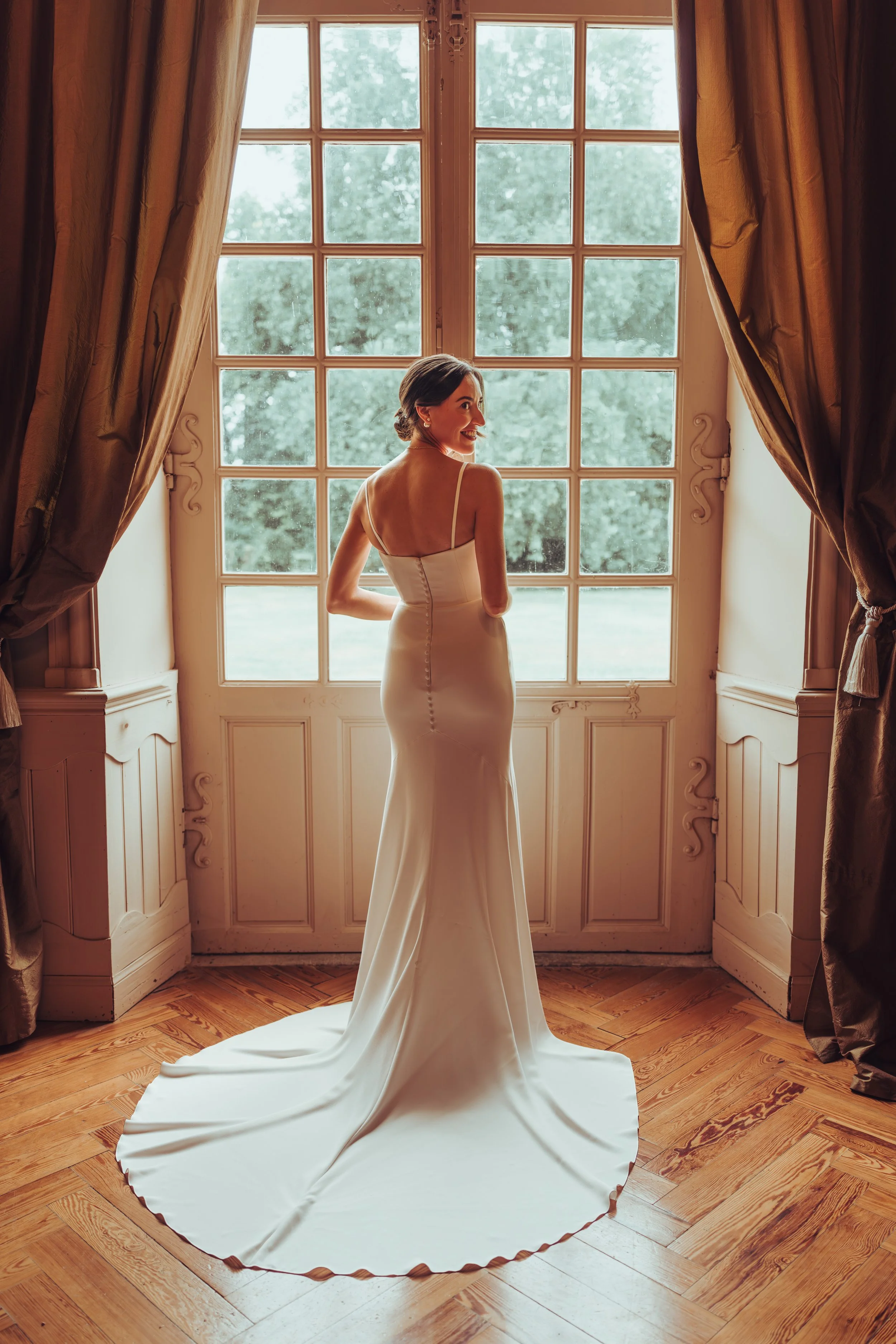Jeune femme en robe de mariée blanche à l'Orangerie des Demoiselles près de Toulouse, en Occitanie regardant par une grande fenêtre avec des rideaux marron, dans une pièce lumineuse avec parquet en bois.