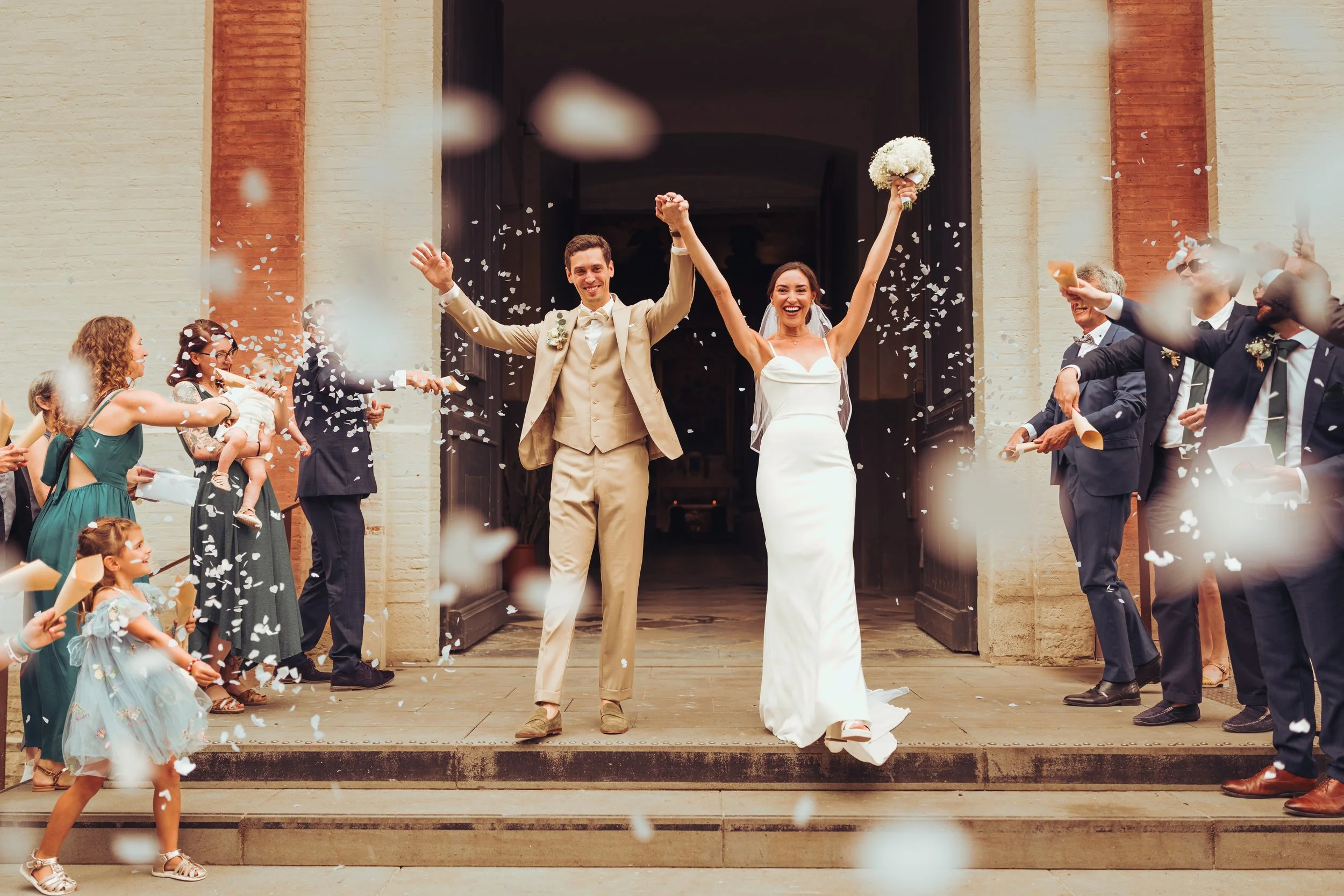 Un couple marié heureux sortant de l'église de Seysse, près de Toulouse, en Occitanie, entouré d'amis et de famille qui leur jettent des confettis, sous des pétales de fleur.