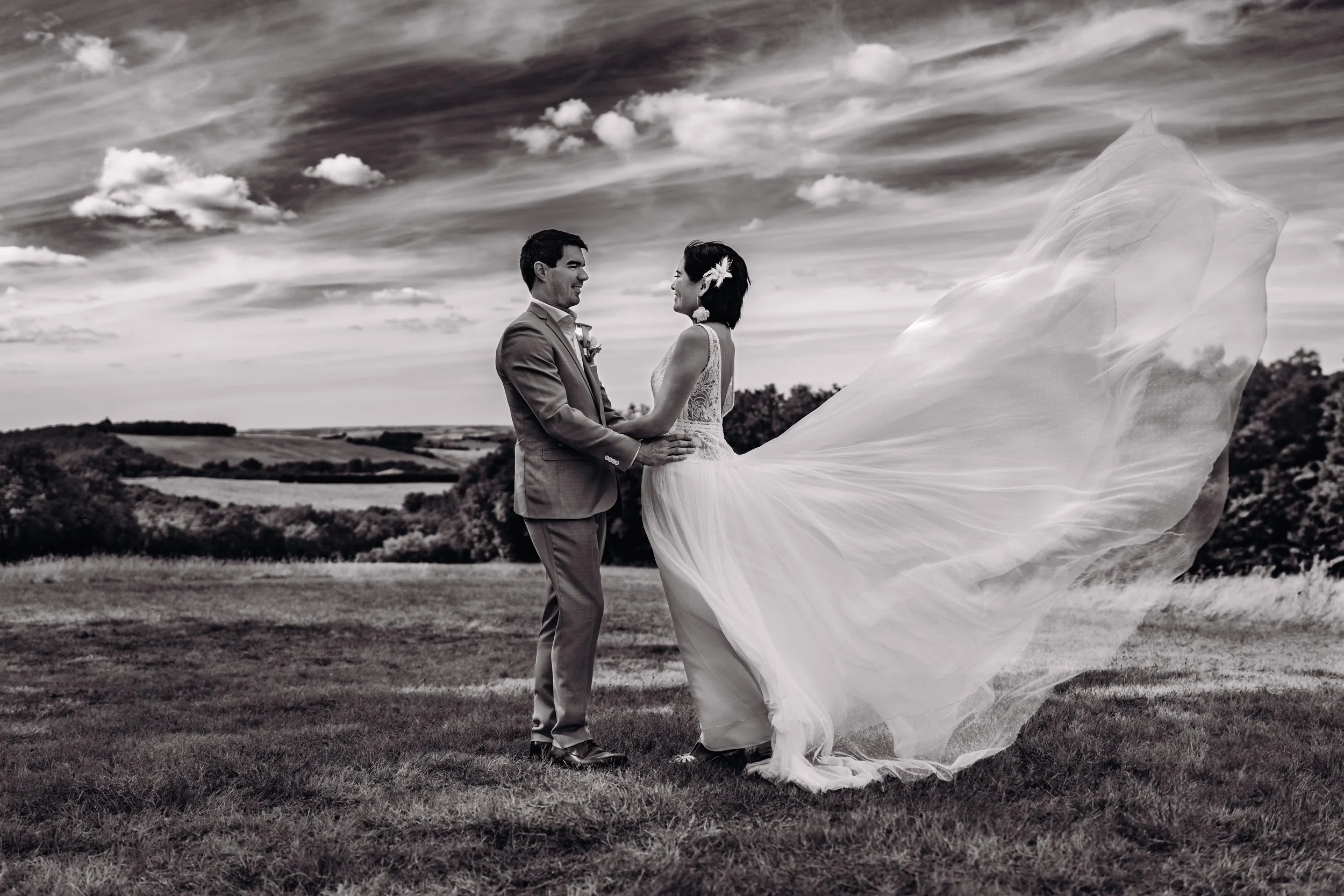 Un couple de mariés, homme en costume et femme en robe de mariée, se tient dans un champ avec un ciel nuageux en arrière-plan. La robe de la mariée flotte dans le vent à la métairie du Fusté près de Toulouse, en Occitanie. 
