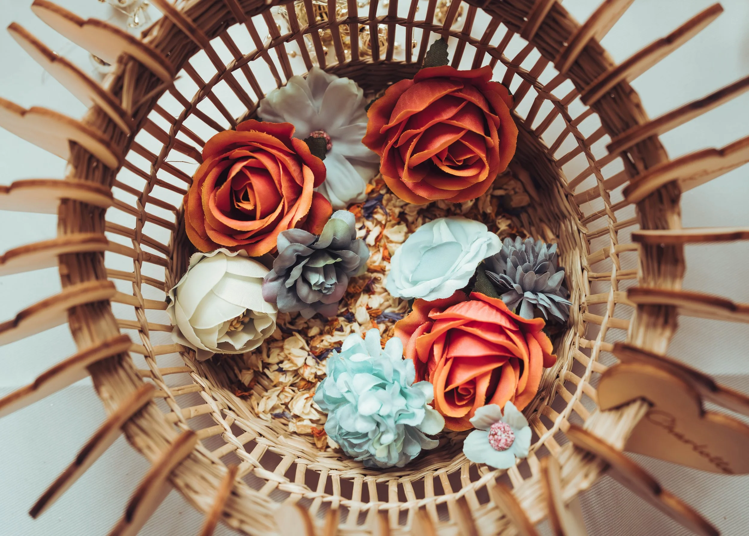 Un panier en rotin rempli de fleurs artificielles de différentes couleurs, y compris des roses orange, des hortensias gris et blancs, et d'autres fleurs avec des pétales colorés.