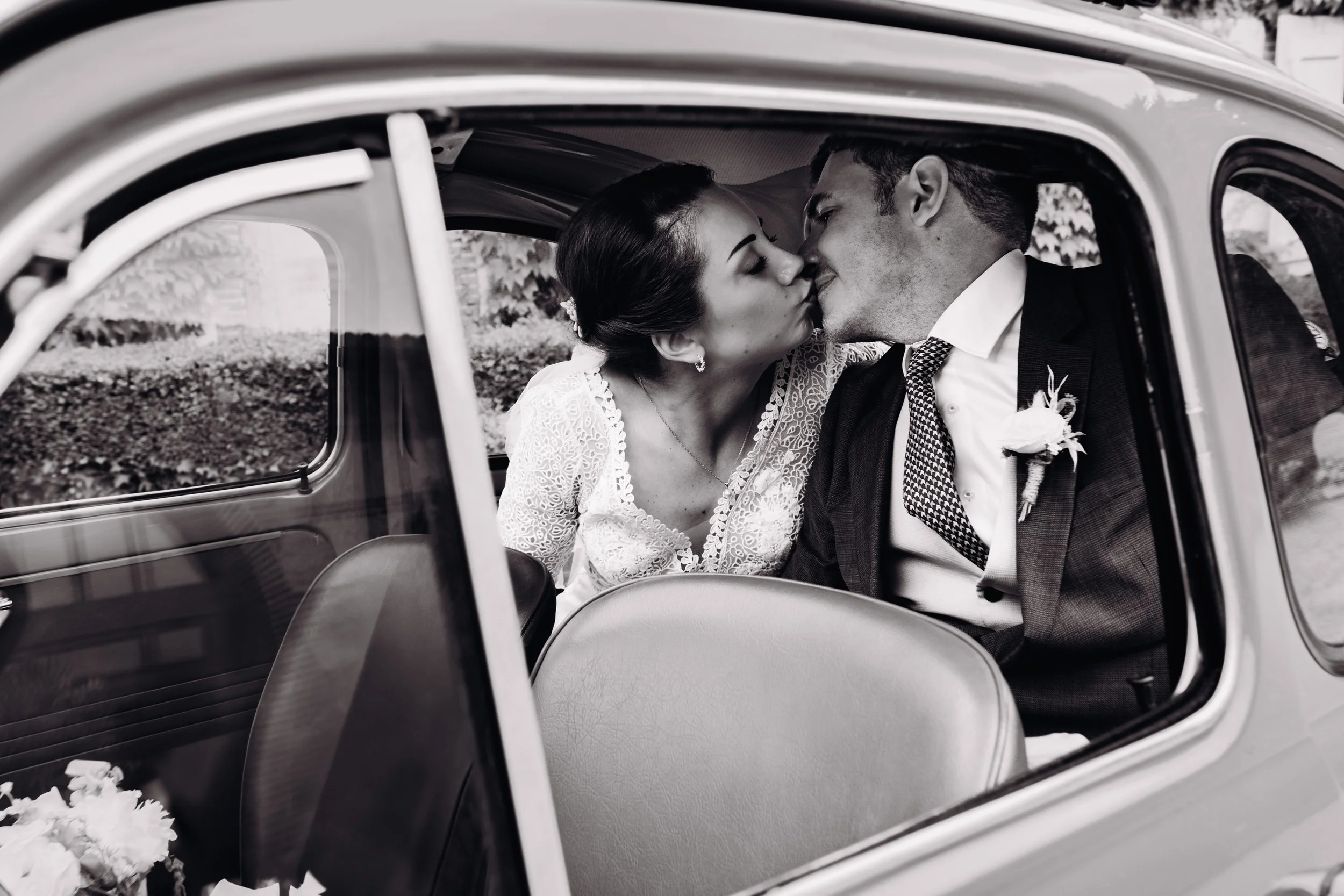 Un couple en mariage s'embrasse dans une voiture ancienne au domaine de Gailhaguet proche de Toulouse, en Occitanie. L'image est en noir et blanc.