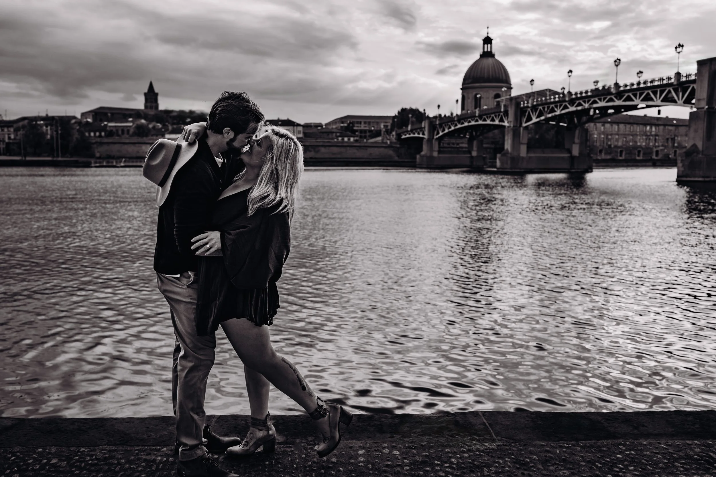 Un couple en pose romantique au bord de la rivière avec un pont et une cathédrale en arrière-plan, en noir et blanc.