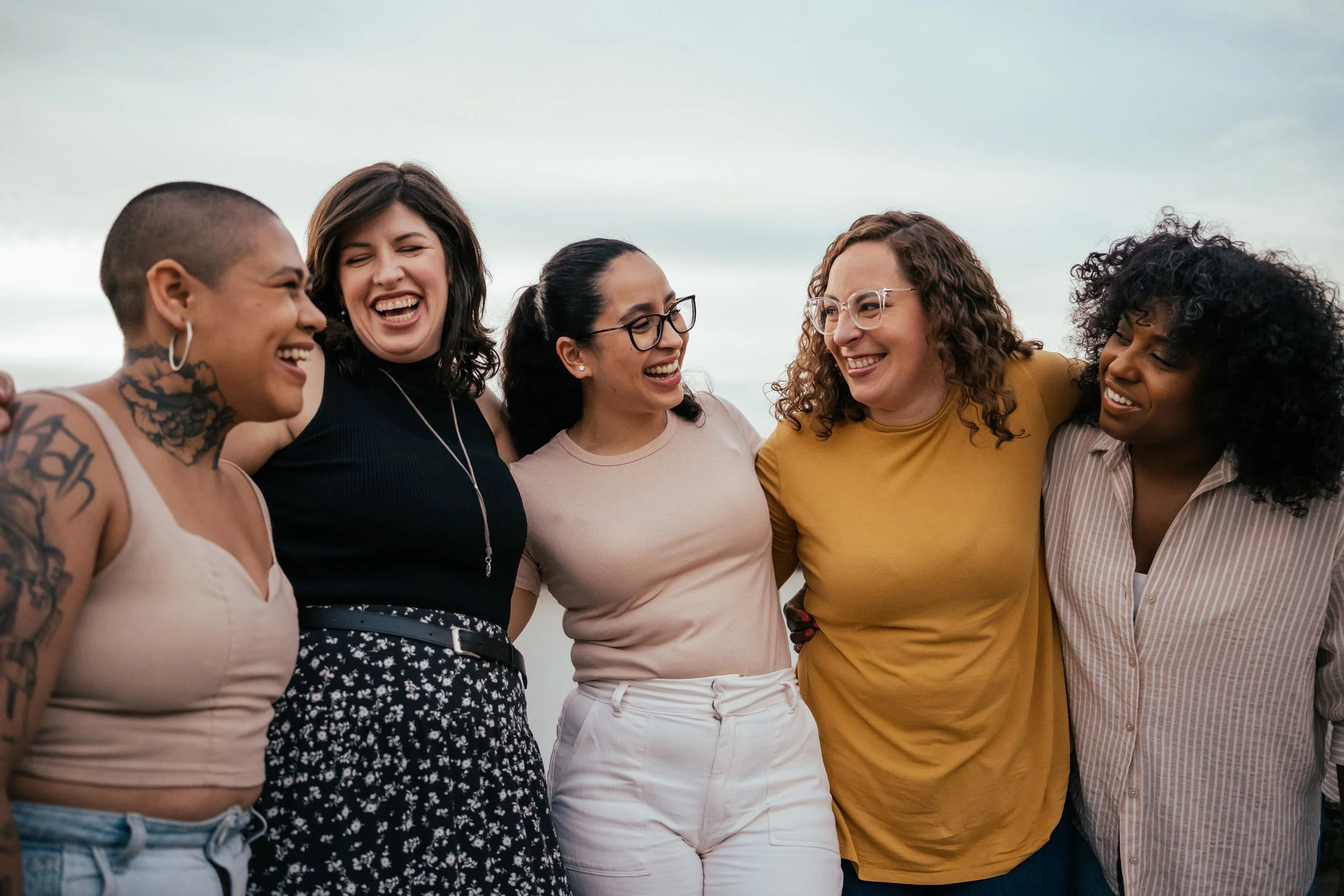 Five women standing close together outdoors, smiling and laughing, with arms around each other, against a cloudy sky.