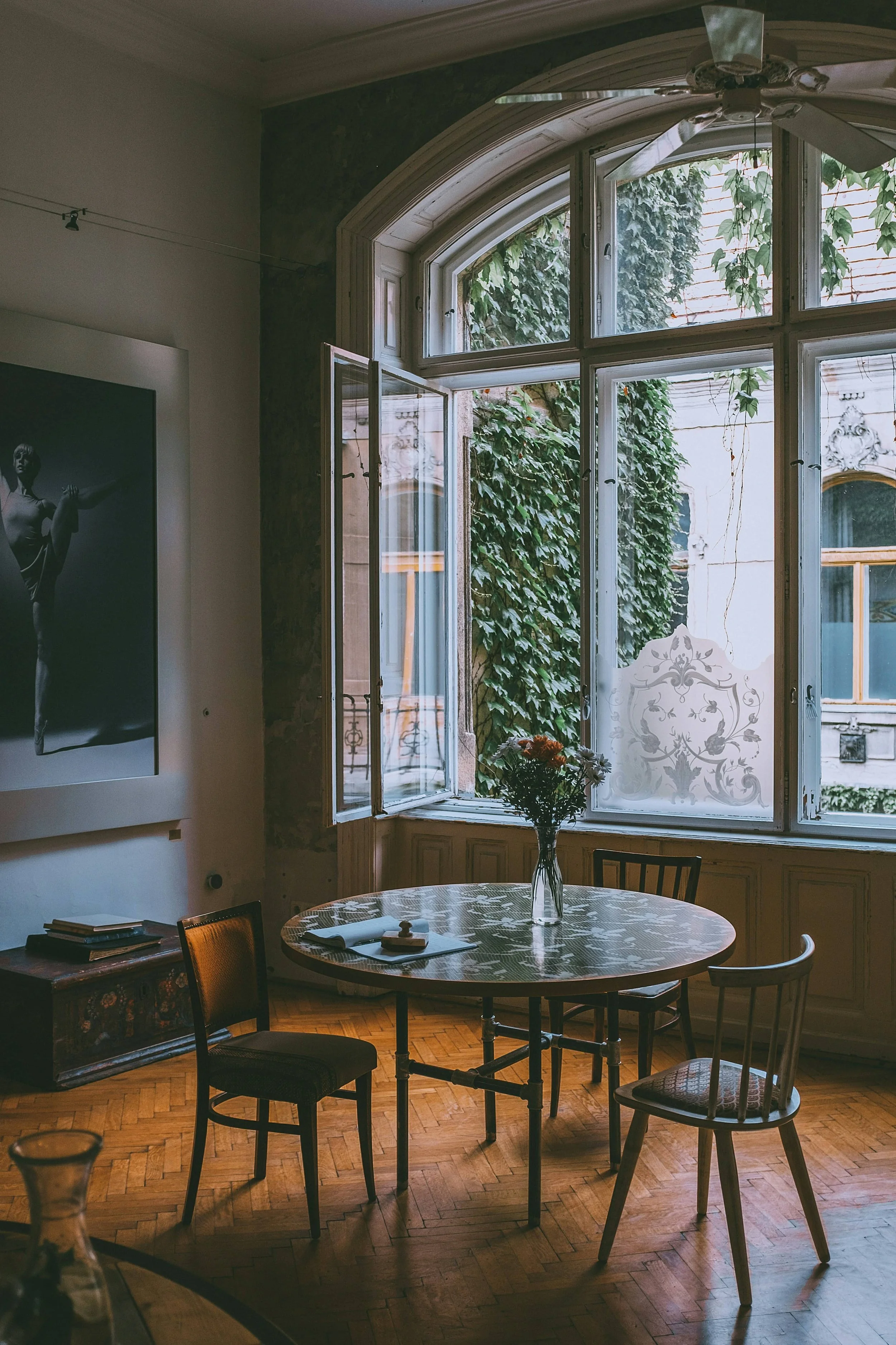 Interior of a room with a round table, a vase of flowers, open window with greenery outside, and a black-and-white statue photo on the wall.