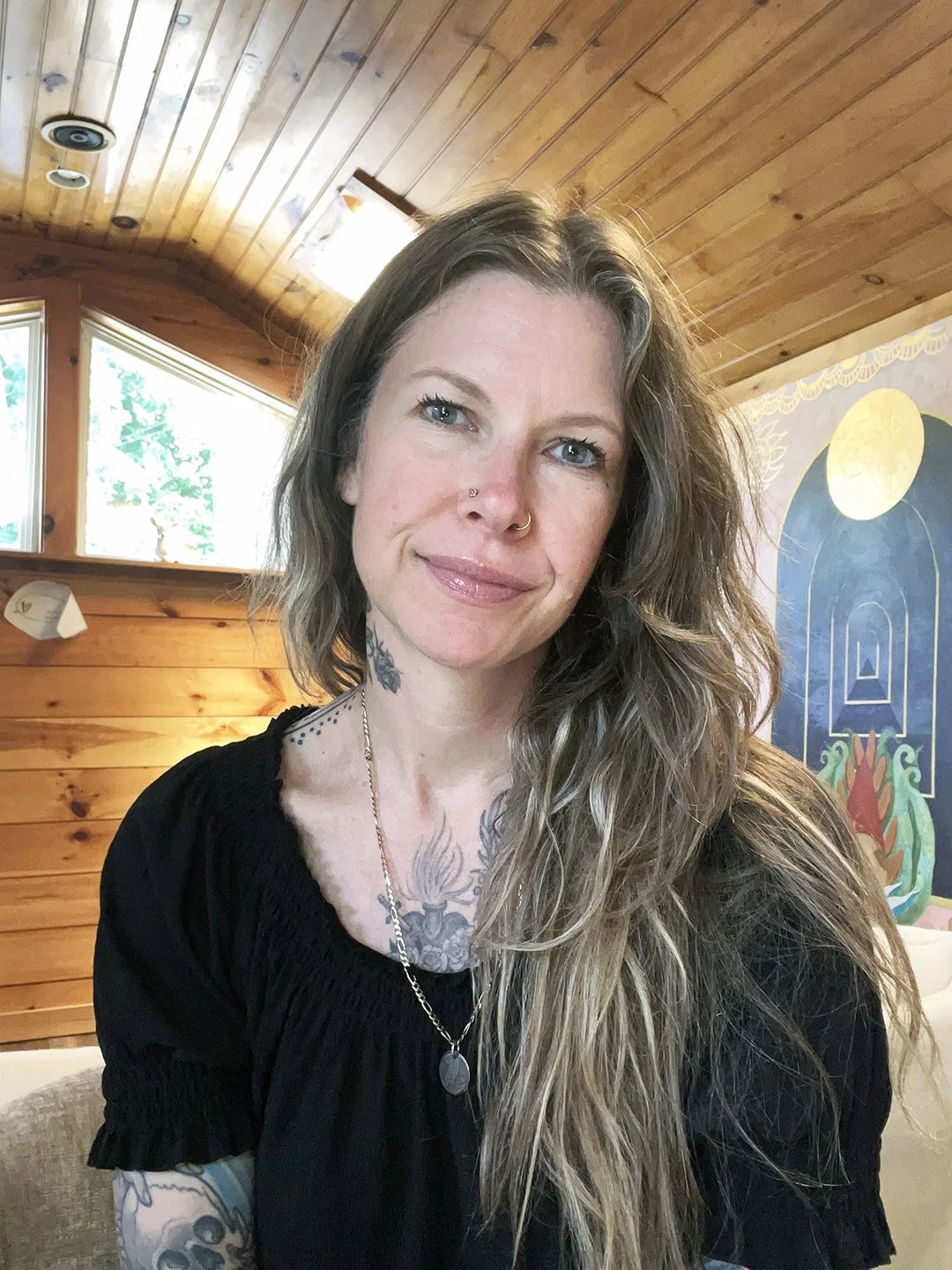 A photo of Dyanna in her space, sitting indoors with wooden walls and a mural of a moon and abstract shapes in the background.