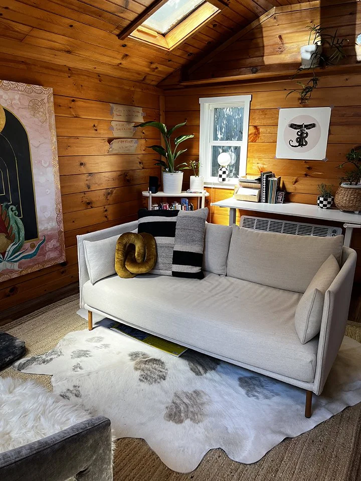 Dyanna’s office, with wooden walls and ceiling, a white couch with pillows, a cowhide rug, a small window, and artwork on the walls. There's a skylight in the ceiling bringing in natural light.