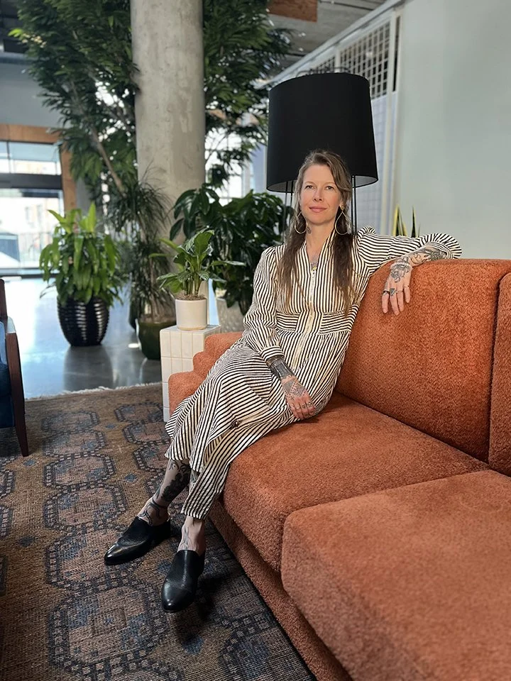 A photos of Dyanna wearing a striped jumpsuit and black shoes, sits on an orange sofa in a modern, plant-filled indoor space.
