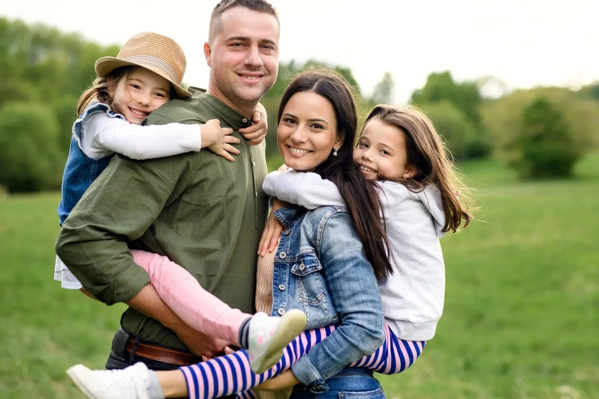 A happy family of four outdoors in a green park. The dad is holding a young girl on his back, and the mom is holding another girl, both smiling and hugging. The family is casually dressed and enjoying sunny weather.
