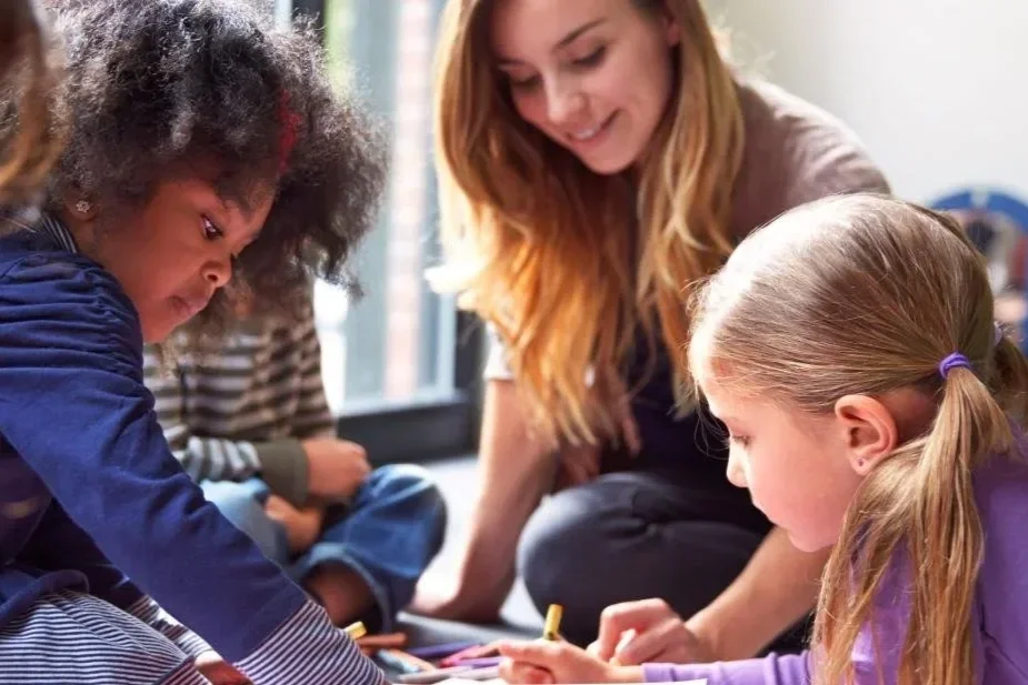 A diverse group of children and a woman sitting on the floor engaged in an activity together, with a window in the background.