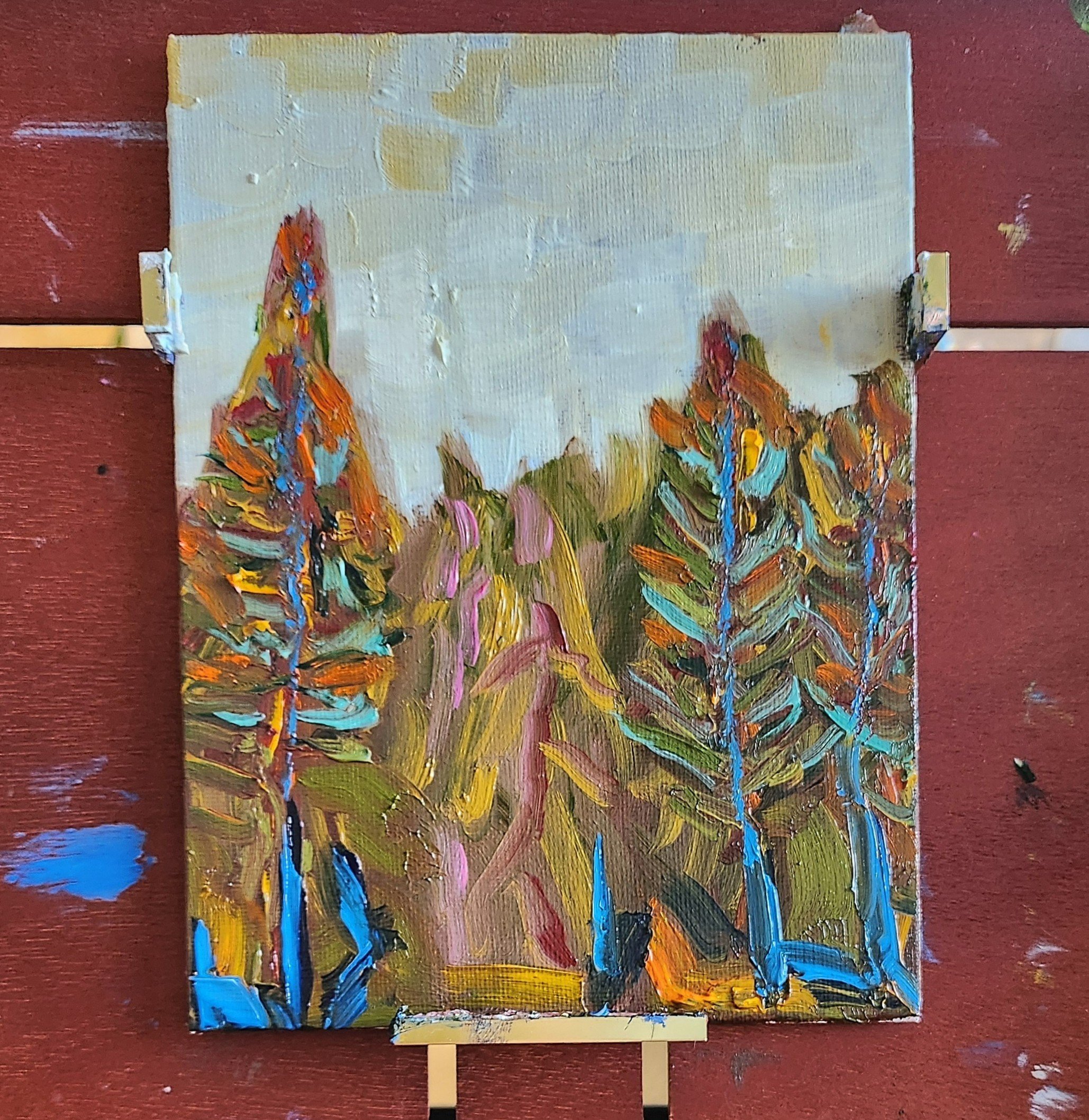An oil painting done in the abstract impressionist style. This landscape painting is of trees, with colorful, textured brush strokes in orange, blue, green, pink, and yellow, arranged on a small canvas on an easel against a red wall.