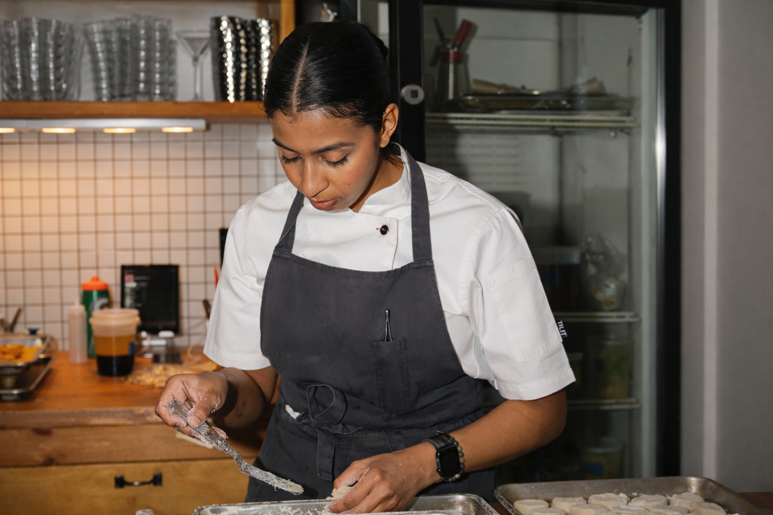 A female chef in a white uniform and black apron preparing food in a kitchen.