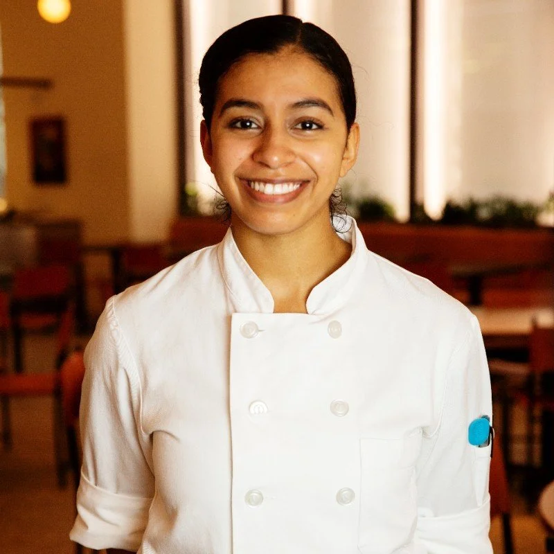 A woman in a white chef's coat smiling in a restaurant setting.