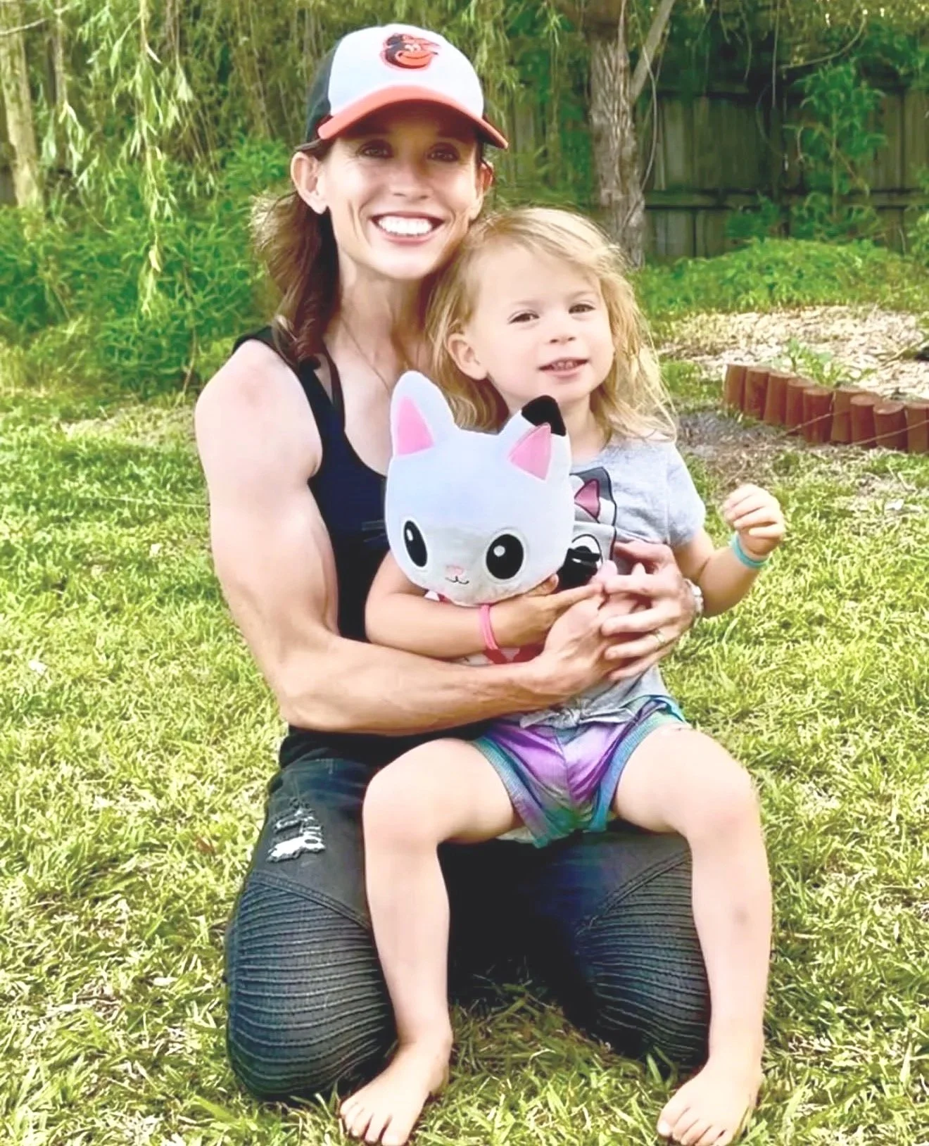 A woman and a young girl sitting on the grass outdoors, smiling at the camera. The woman is wearing a baseball cap and a black tank top, holding a plush toy bunny.