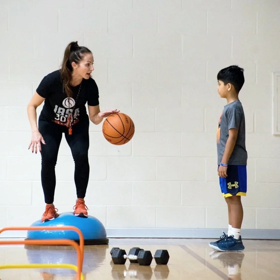 A female coach standing on a blue balance trainer, holding a basketball and talking to a young boy standing on the gym floor. The scene appears to be in a gym or sports practice setting, with weights in the foreground and a plain white wall in the ba