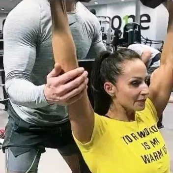 A woman with a ponytail performing a shoulder press exercise with dumbbells at the gym, supervised by a male trainer.
