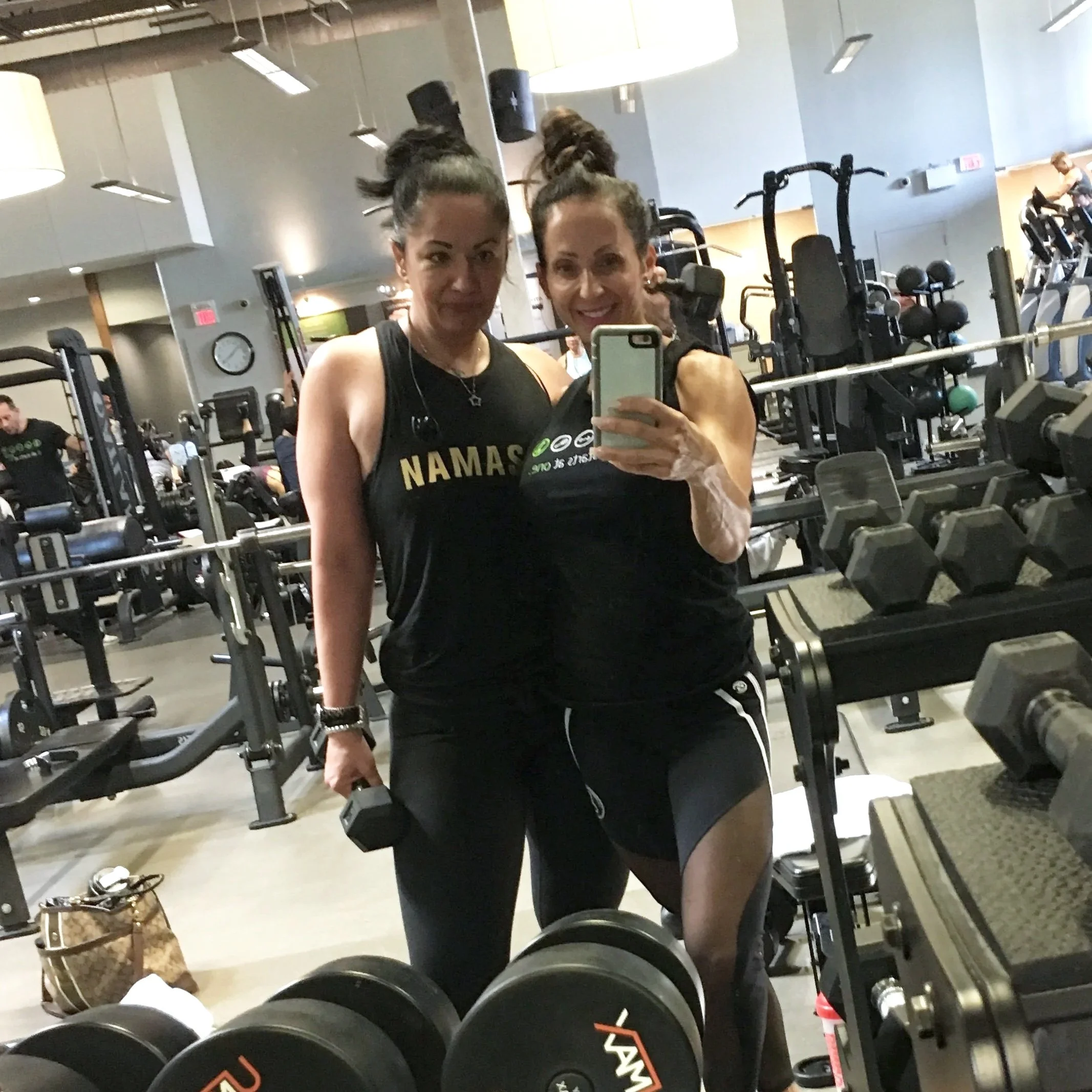 Two women taking a selfie in a gym, one holding a dumbbell, surrounded by gym equipment including dumbbells, weights, and exercise machines.