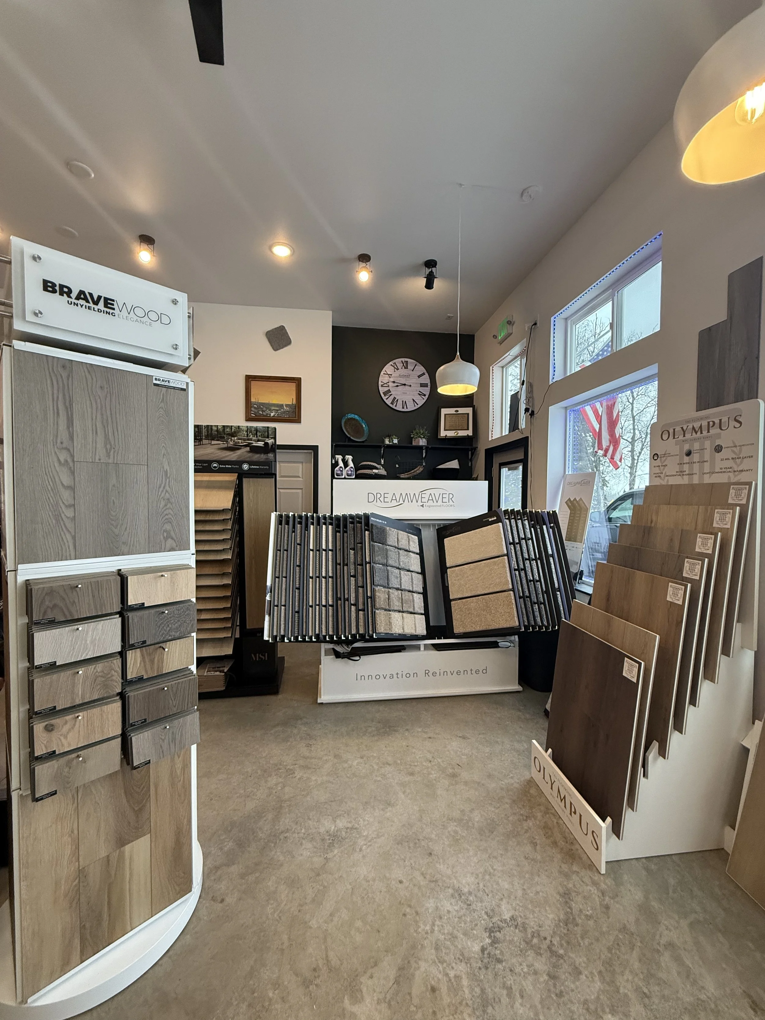 Interior of a flooring store displaying samples of wood, tile, and laminate flooring. There are sample display racks, a large window, and a clock on the back wall.