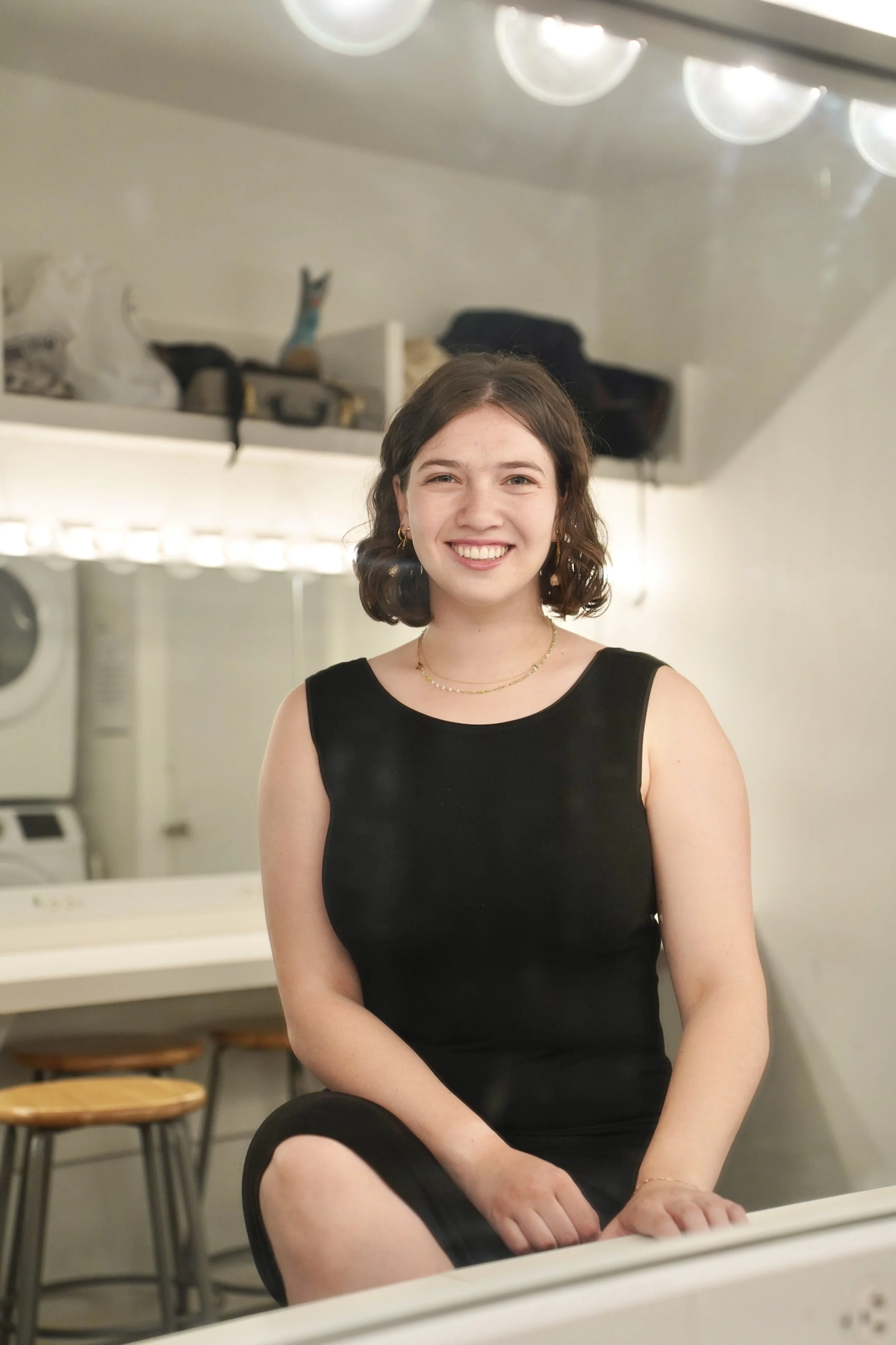 A young woman with brown, shoulder-length hair smiling at the camera, wearing a black sleeveless top and gold jewelry, in a room with washing machines and chairs in the background.