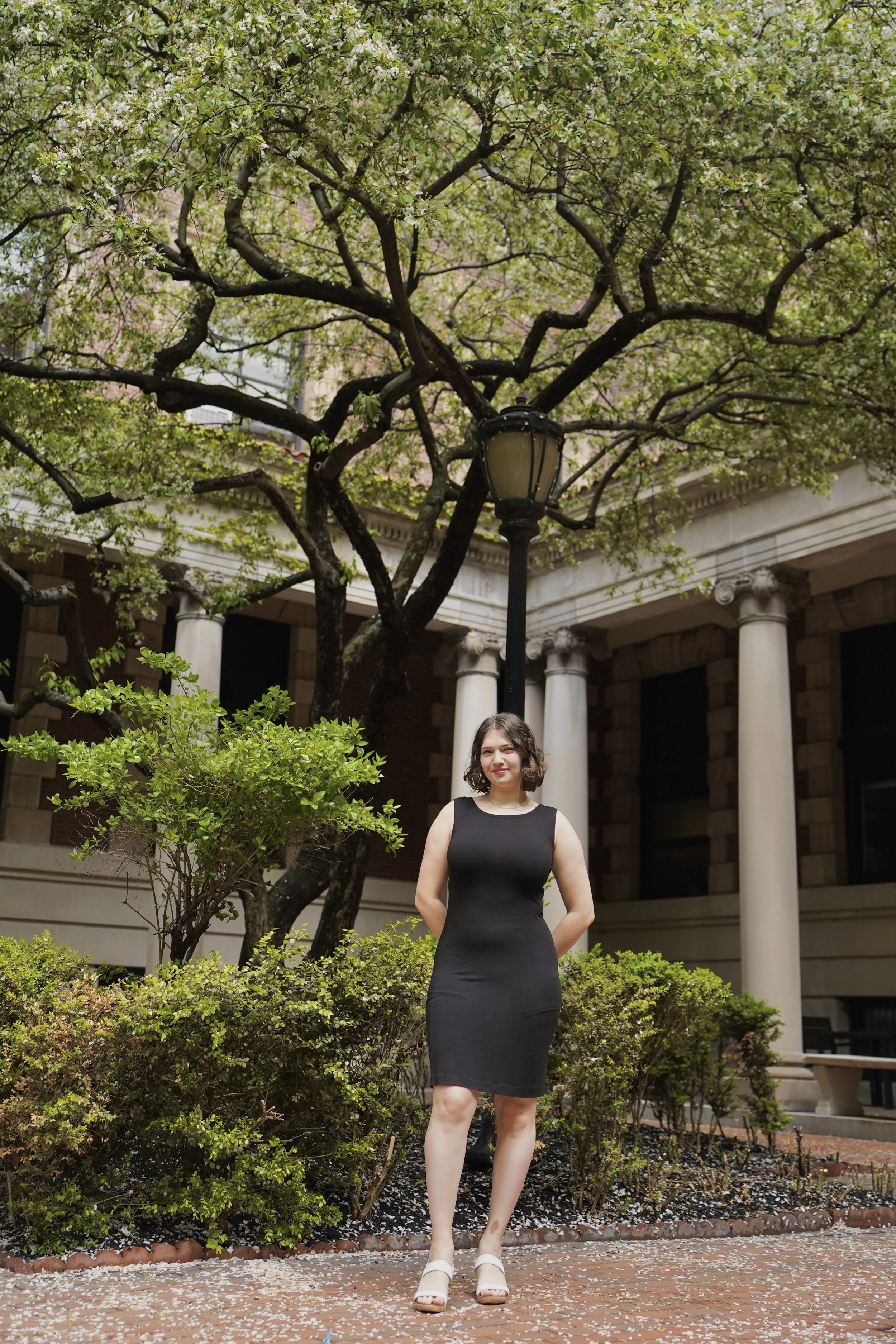 A woman in a fitted black dress standing outdoors in front of a building with columns and surrounded by greenery, including trees and bushes.
