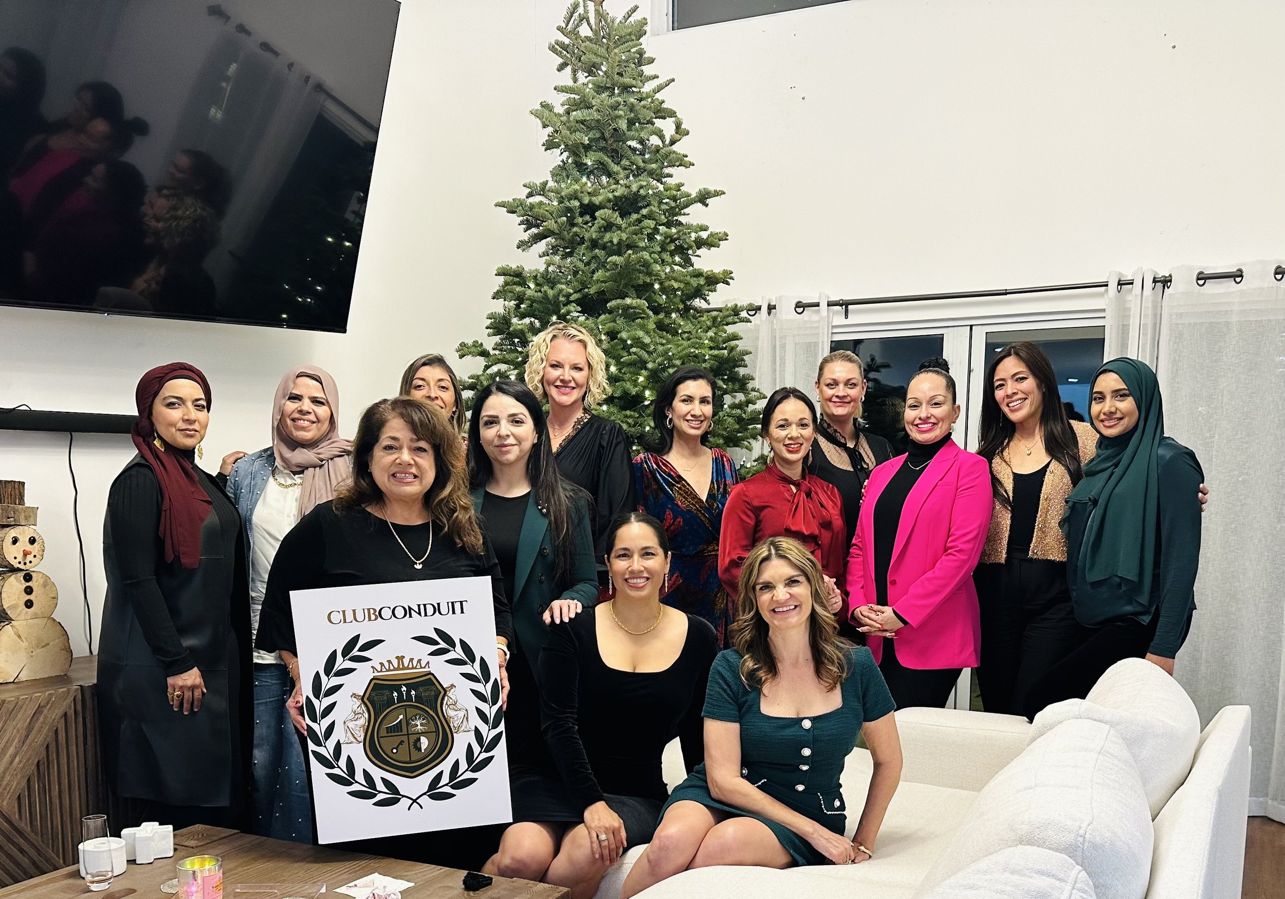 A group of women gathered around a Christmas tree in a living room, posing for a photo. There is a large TV on the wall and a sign reading 'Club Conduit' with a logo. The women are smiling and dressed in various colorful outfits.