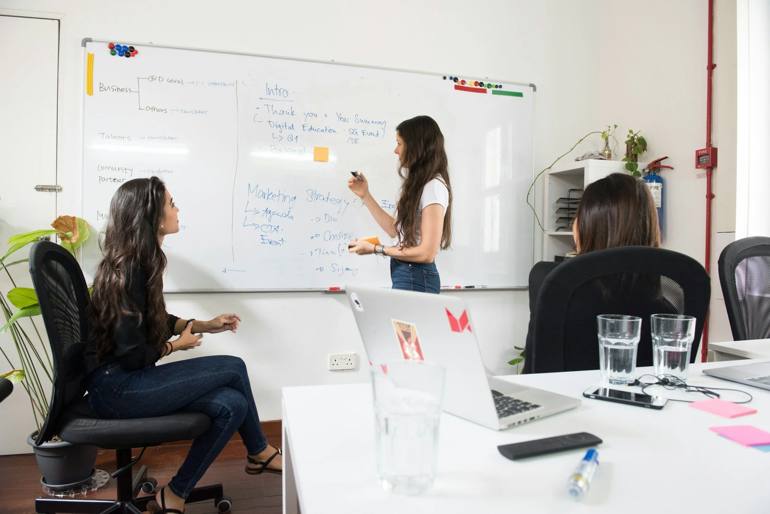 A woman standing and writing on a whiteboard while three women sit and listen in a modern office conference room with laptops, water glasses, and plants.