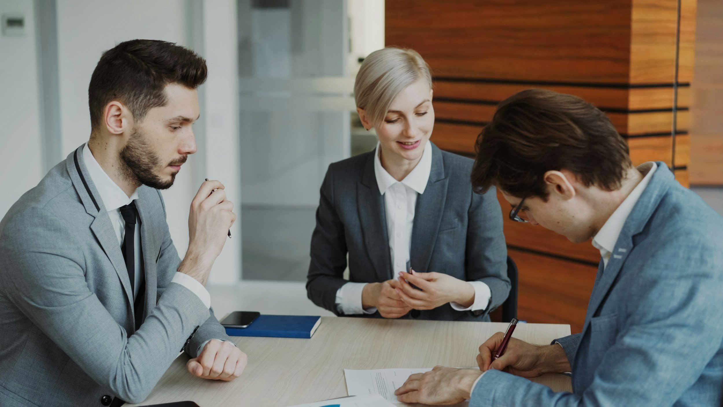 Three business professionals, two men and one woman, in a meeting room, reviewing documents and discussing work.
