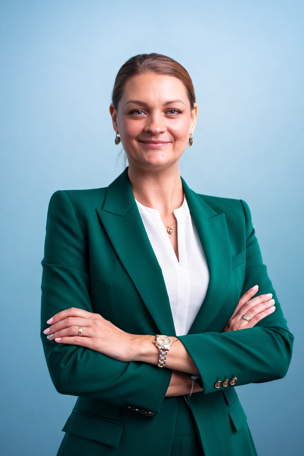 A woman in a green blazer with arms crossed, smiling, against a light blue background.