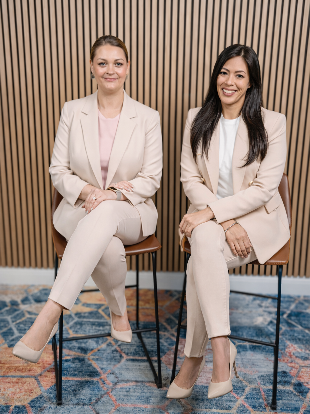 Two women sitting on chairs in front of a wooden slat wall, dressed in beige suits and high heels, smiling at the camera.