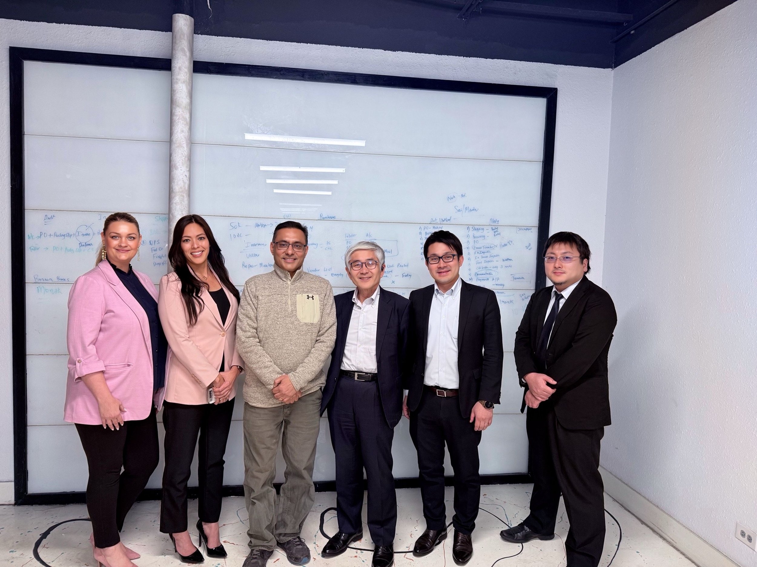 Group of six diverse professionals standing together in front of a whiteboard with notes, in a conference room.