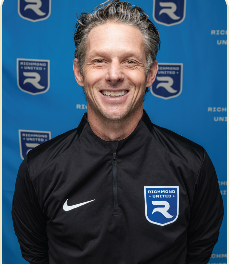 A smiling man wearing a black Nike sports jacket with the Richmond United logo, standing in front of a blue Richmond United backdrop.