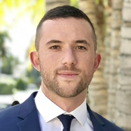 A young man with short dark hair and a beard, wearing a navy suit and white shirt, standing outdoors with a blurred background of trees and a stone wall.