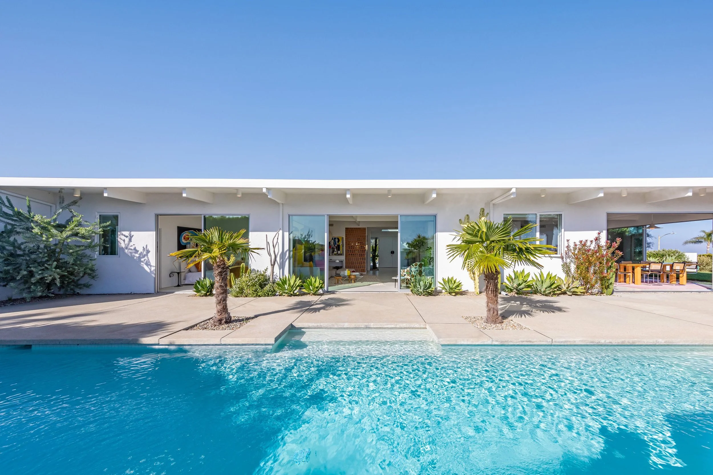 Modern white house with large sliding glass doors, surrounded by desert plants and palm trees, with a swimming pool in the foreground on a clear sunny day.