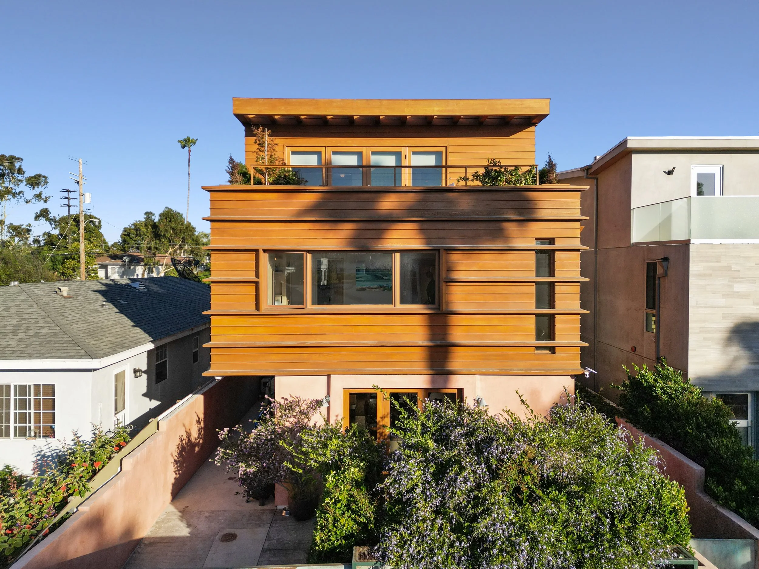 A modern multi-story house with a wooden facade, large windows, and a rooftop terrace, surrounded by lush greenery and neighboring houses, under a clear blue sky.