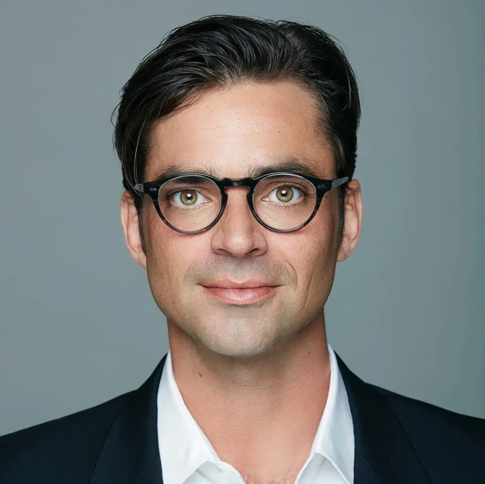 A professional headshot of a man with dark hair, glasses, wearing a black suit and white shirt, against a plain gray background.