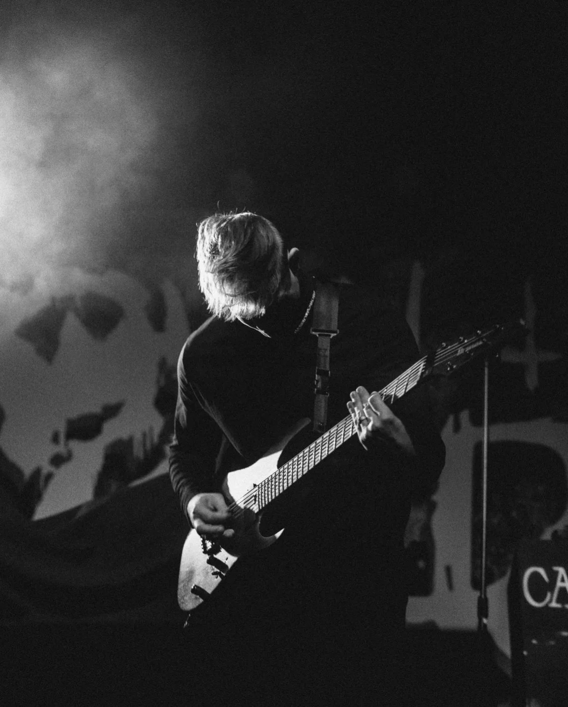 Black and white photo of a person playing an electric guitar on stage, with their head down and hair covering part of their face, likely during a live concert.